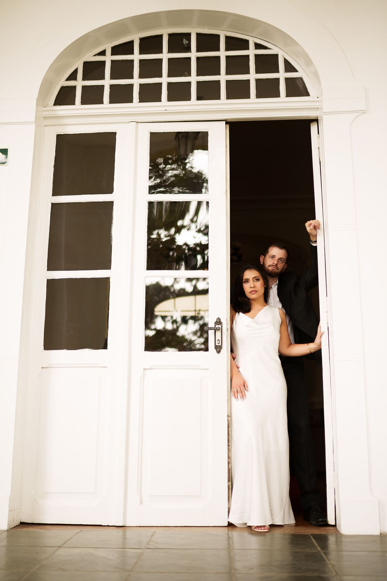 Bride in white gown and groom in black suit posing elegantly in an arched white doorway.