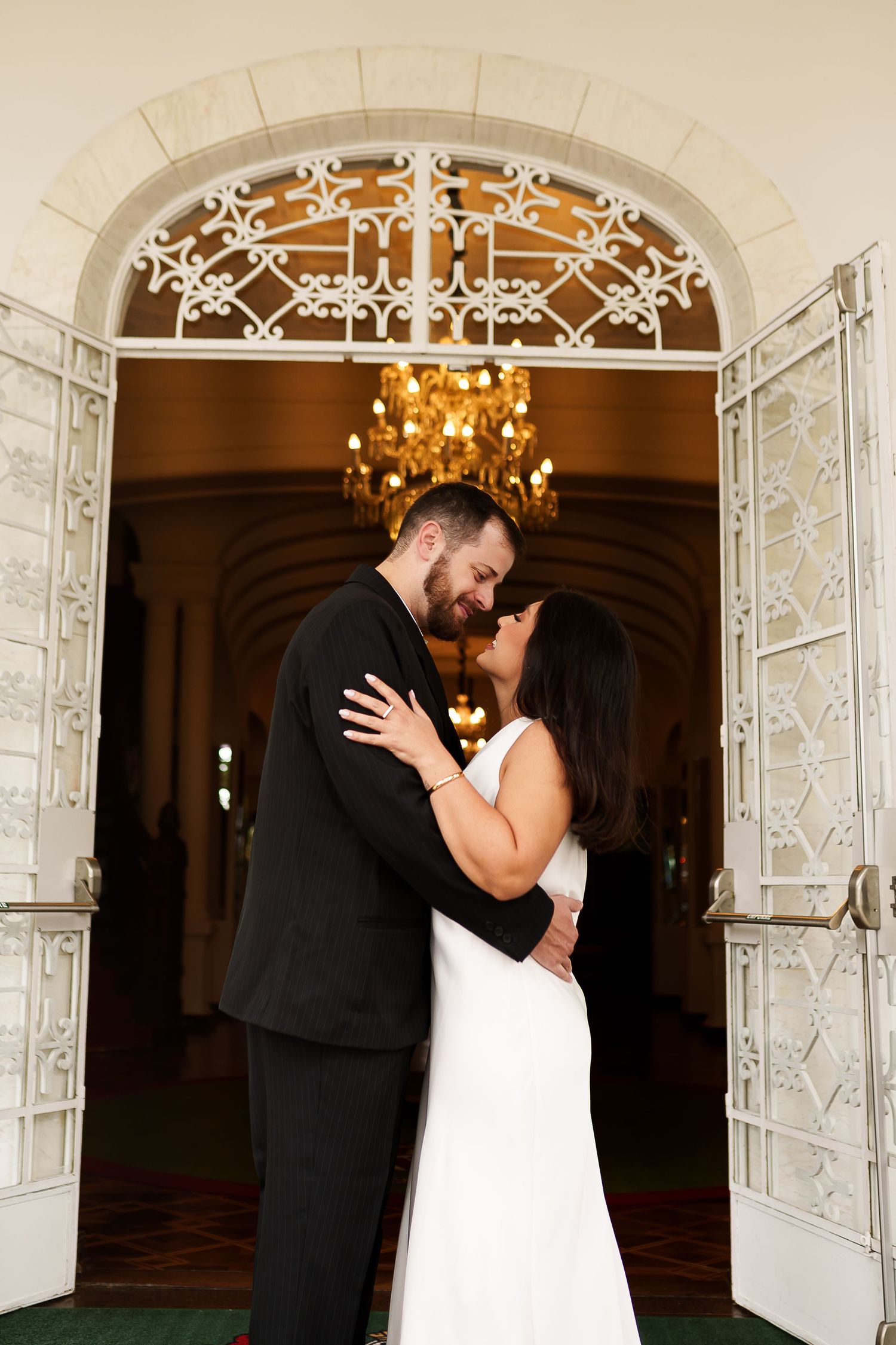 Bride and groom share a romantic kiss in wedding attire beneath an ornate archway with chandelier.
