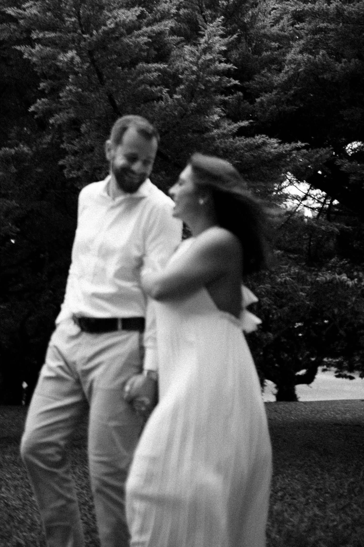 Black and white photo of a smiling couple holding hands and walking together in a lush green park setting.