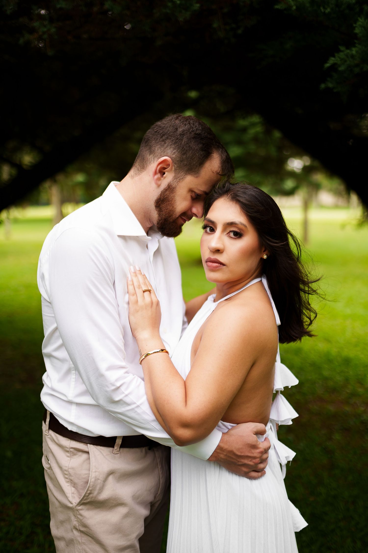 Couple in white outfits embracing in a lush green park, woman looking at camera during romantic engagement photo session.