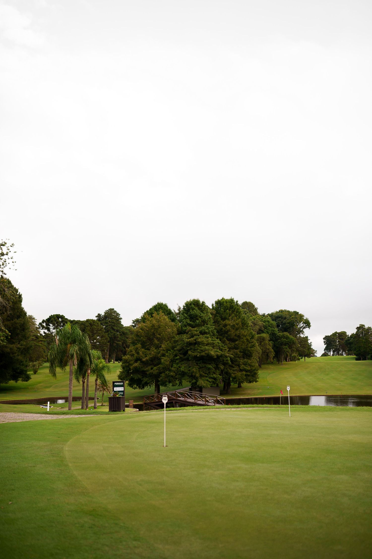 Lush green golf course with white flags, pond, and tree-lined hills under an overcast sky.