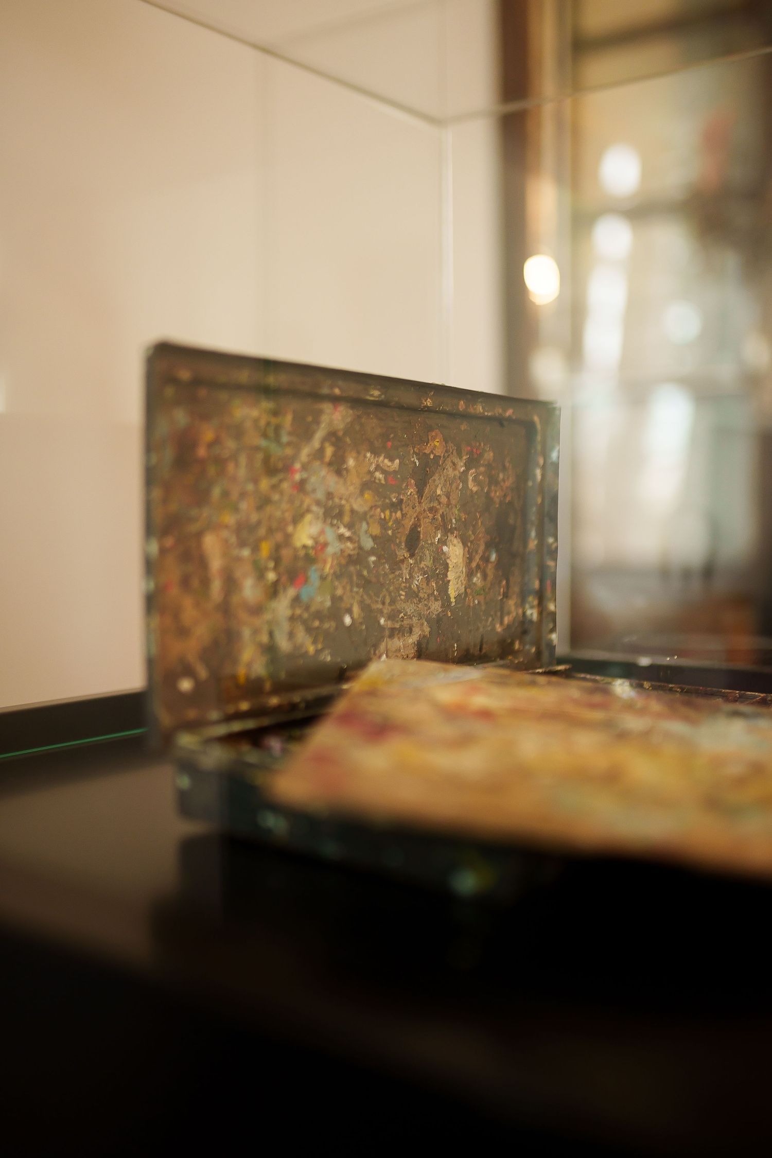 Close-up of paint-splattered artist palettes displayed on a dark shelf in a gallery setting.