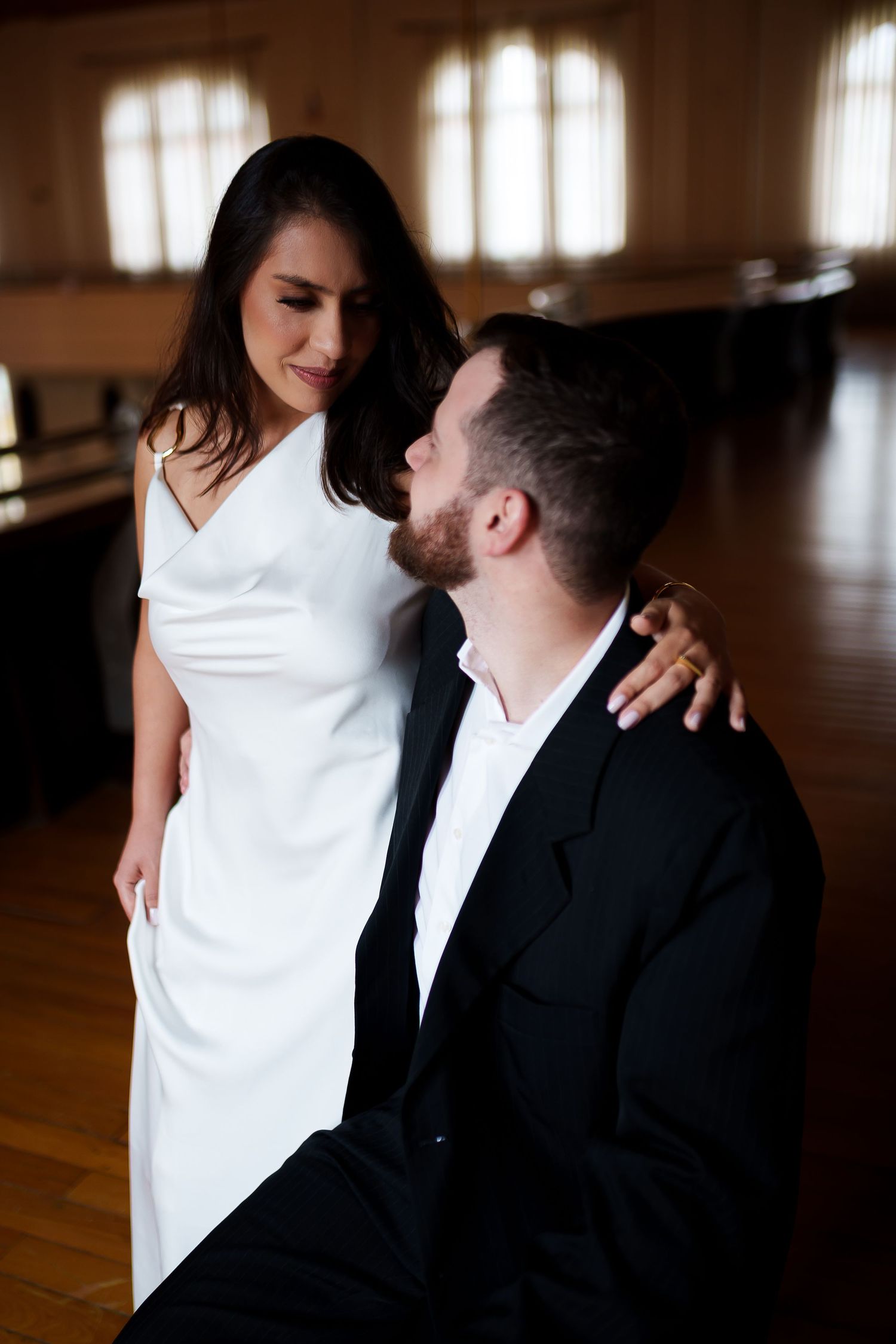 Elegant couple in formal attire sharing a romantic moment, woman in white dress standing beside seated man in black suit.