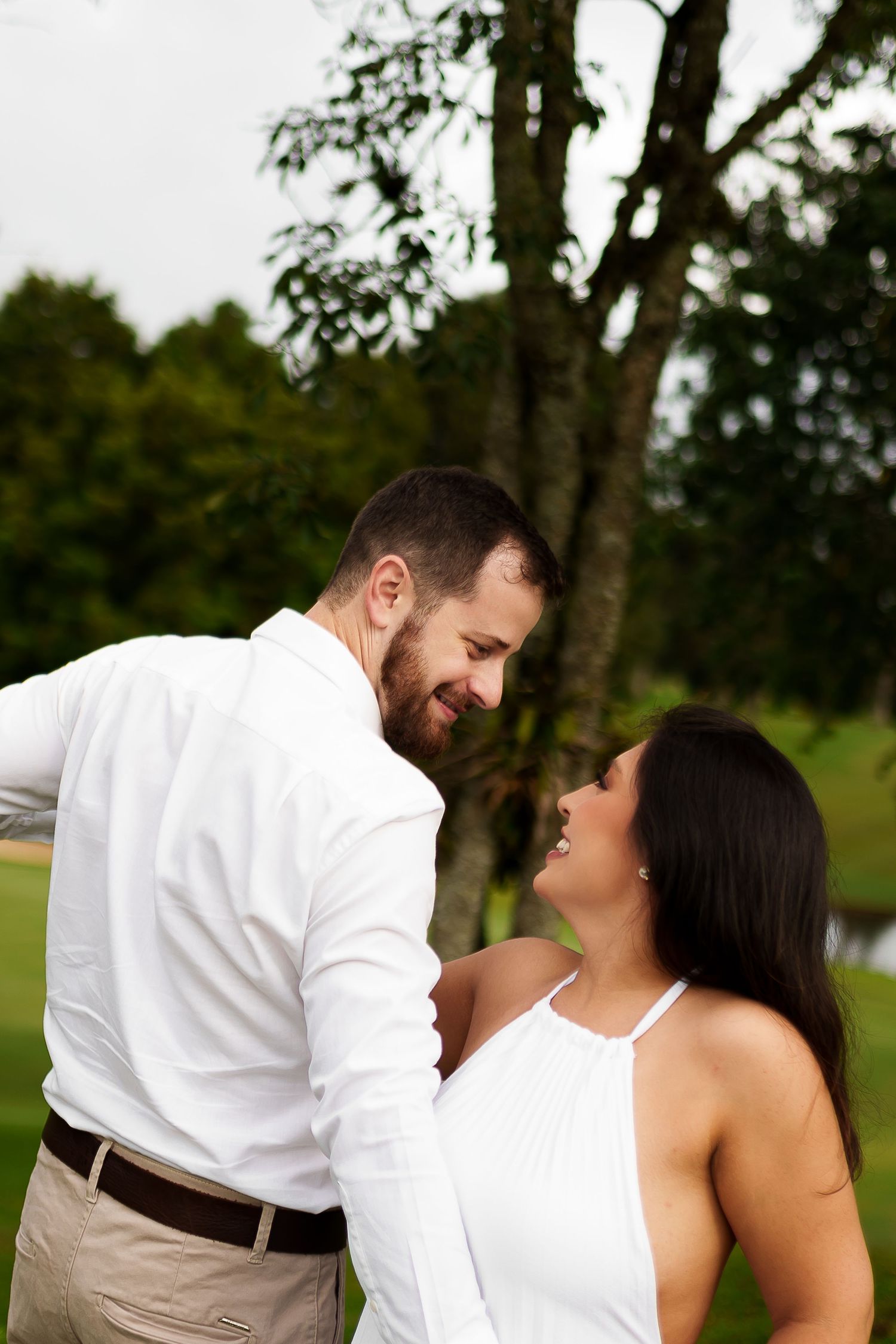 Couple in white outfits sharing a romantic moment outdoors, smiling at each other near a large tree.