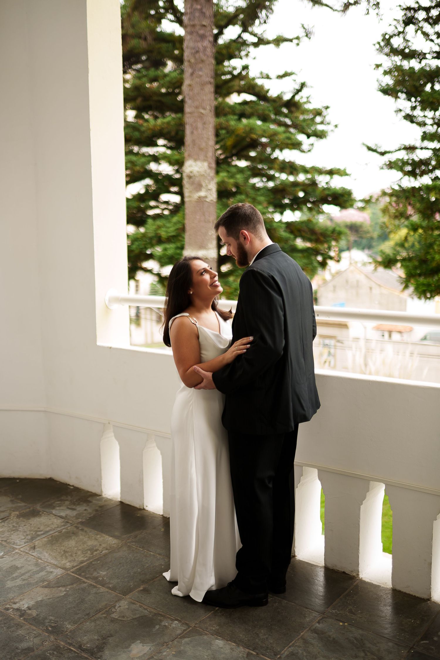 Bride in white dress and groom in black suit share a romantic moment on an elegant white balcony with trees.