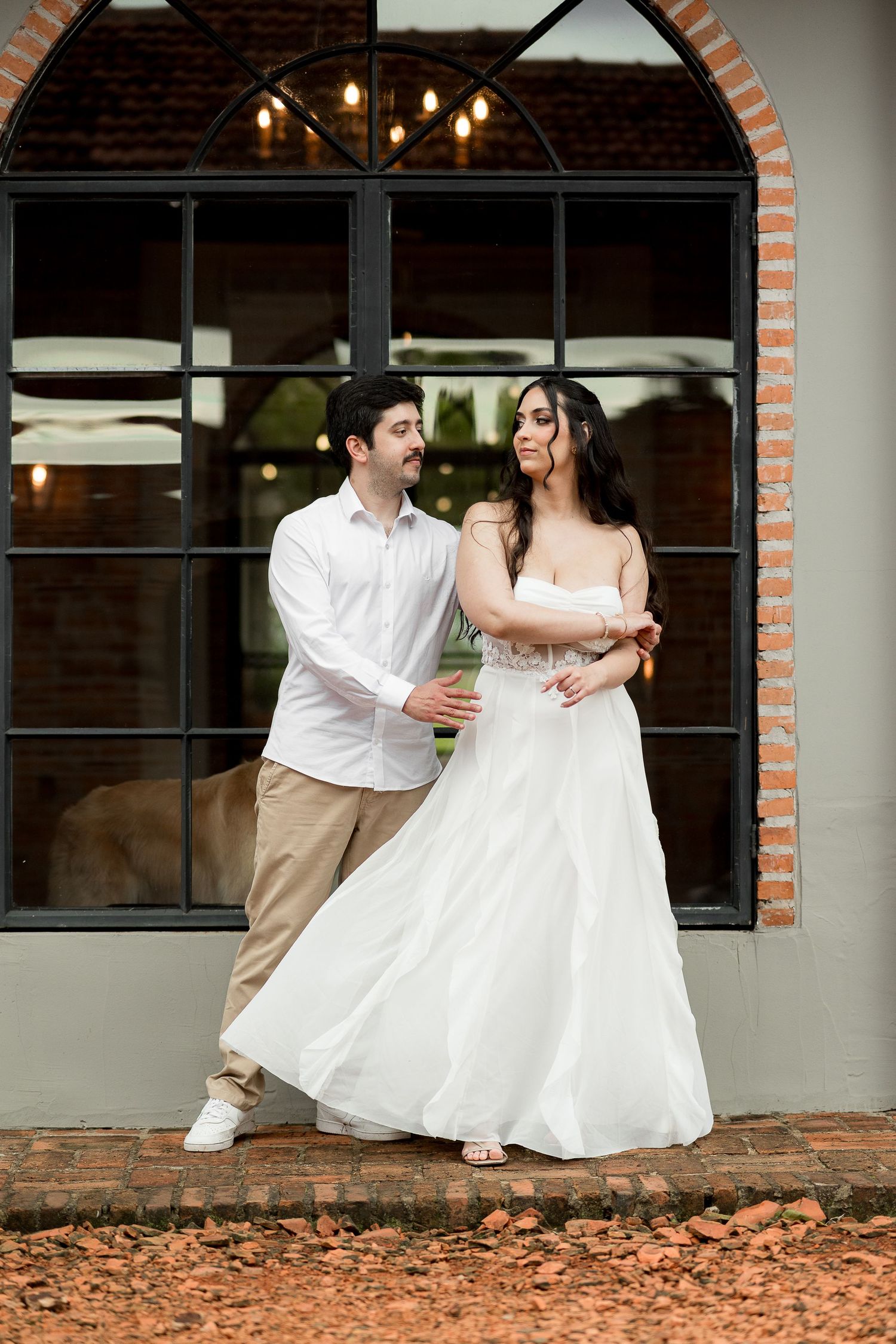 A couple in wedding attire dancing outdoors, bride in white gown and groom in white shirt, posing before an arched window.