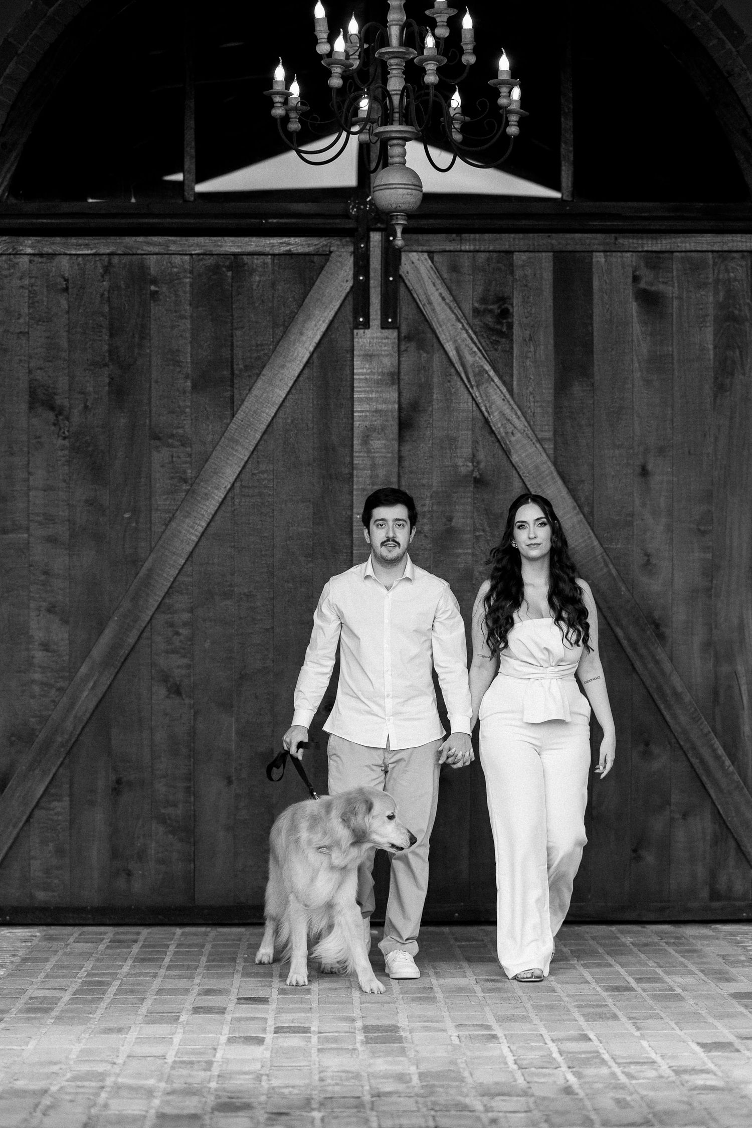 Black and white photo of a couple holding hands with a golden retriever dog in front of a rustic barn door with chandelier.
