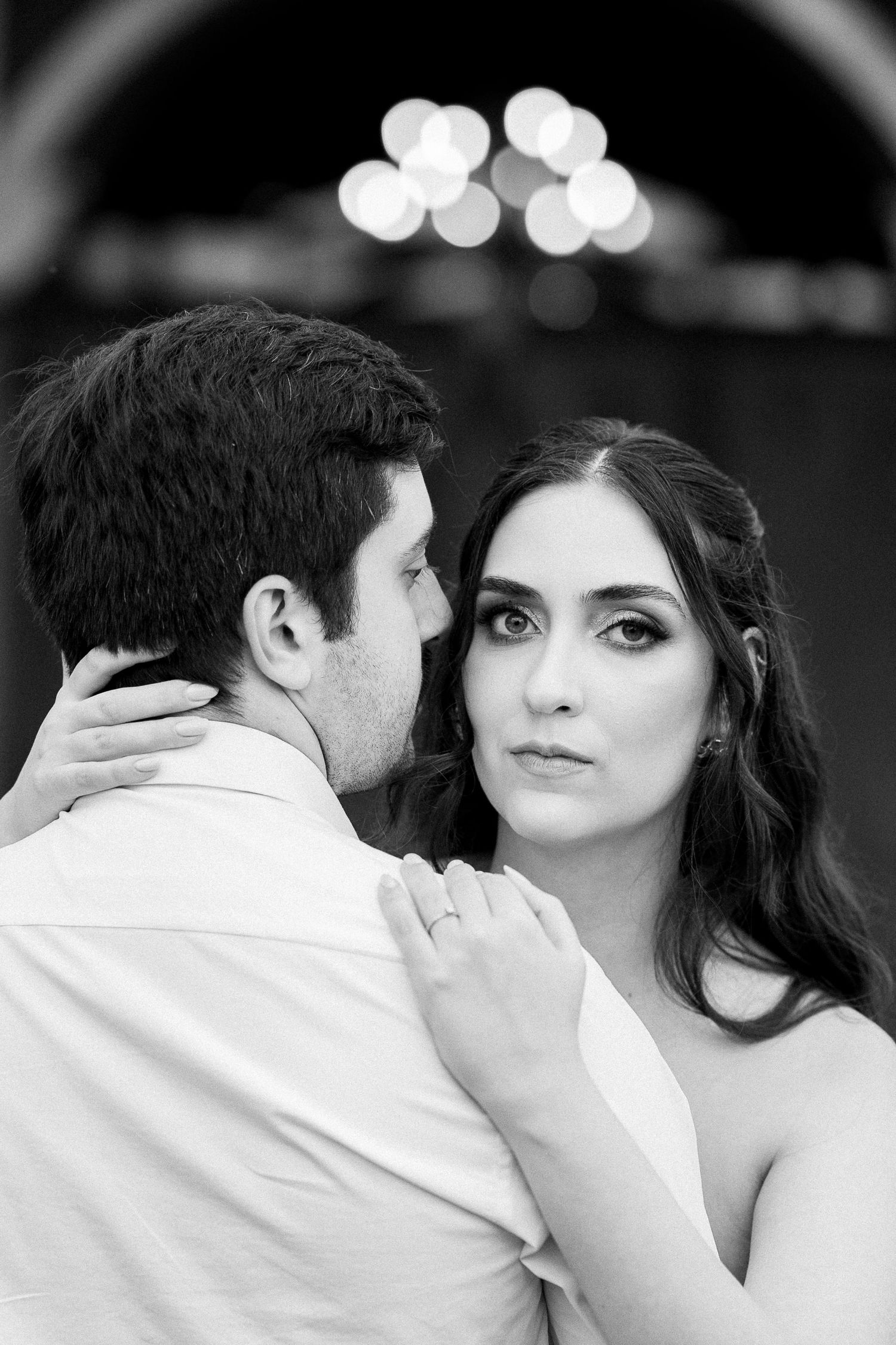 Black and white wedding portrait of bride and groom embracing, bride gazing at camera with bokeh lights in background.