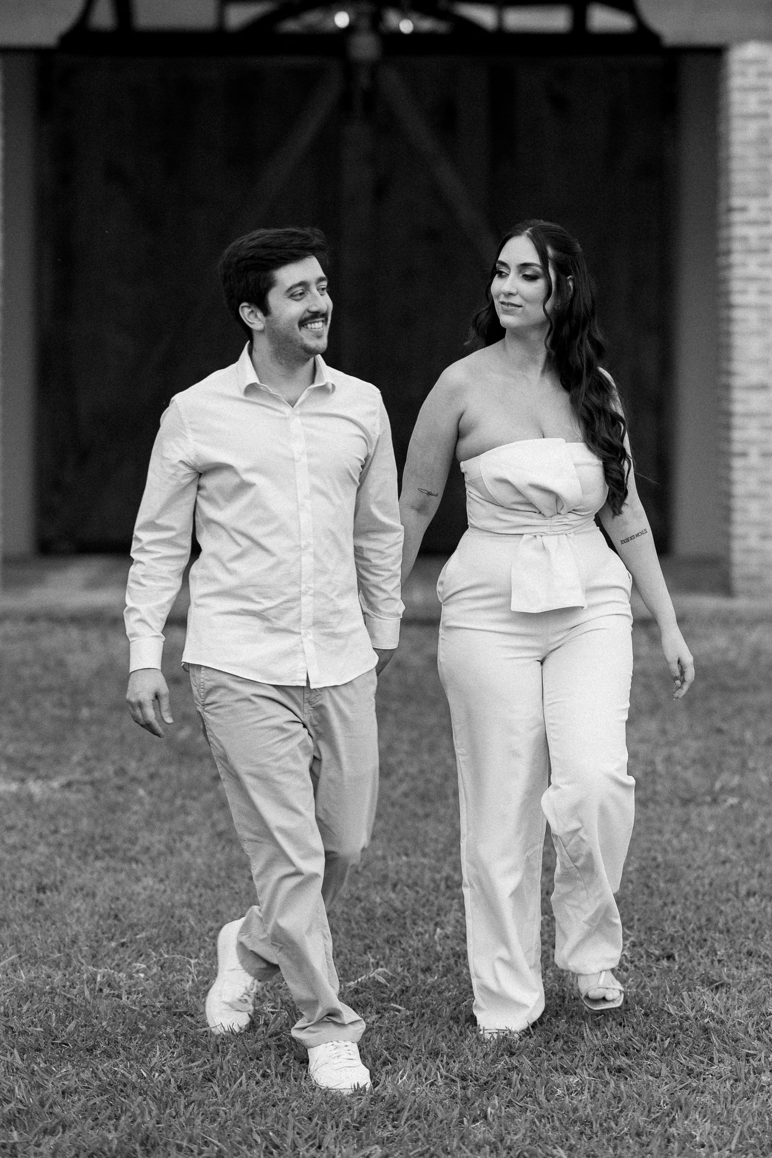 Black and white photo of a smiling couple holding hands and walking on grass in front of a barn.