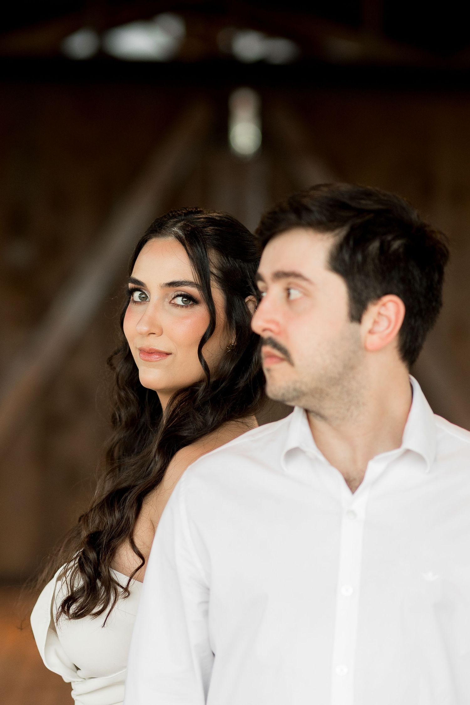 A couple poses romantically in a barn, woman with dark wavy hair in white dress, man in white shirt with mustache.