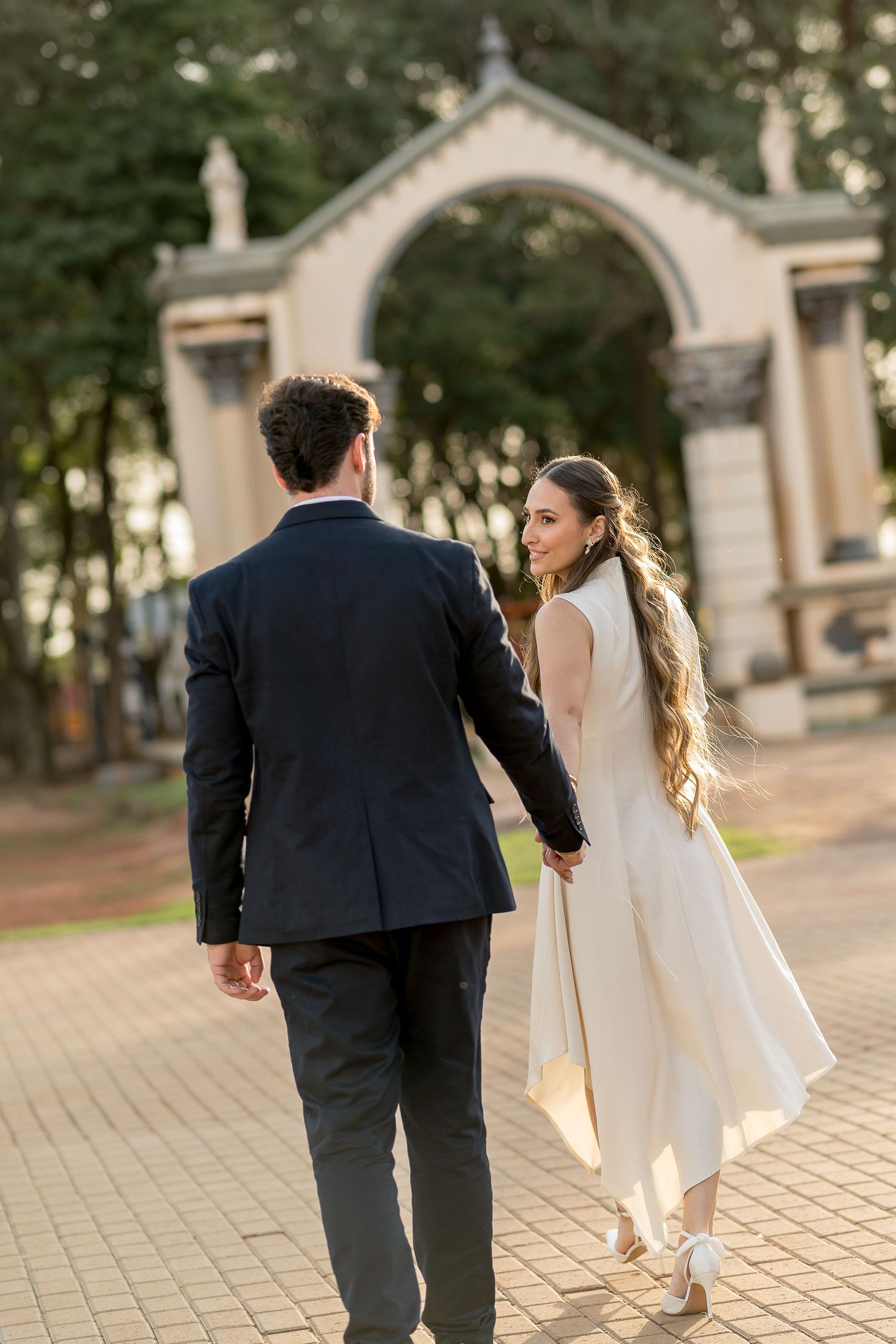 Bride in white dress and groom in navy suit walk hand-in-hand toward an elegant arch in a sunlit garden.