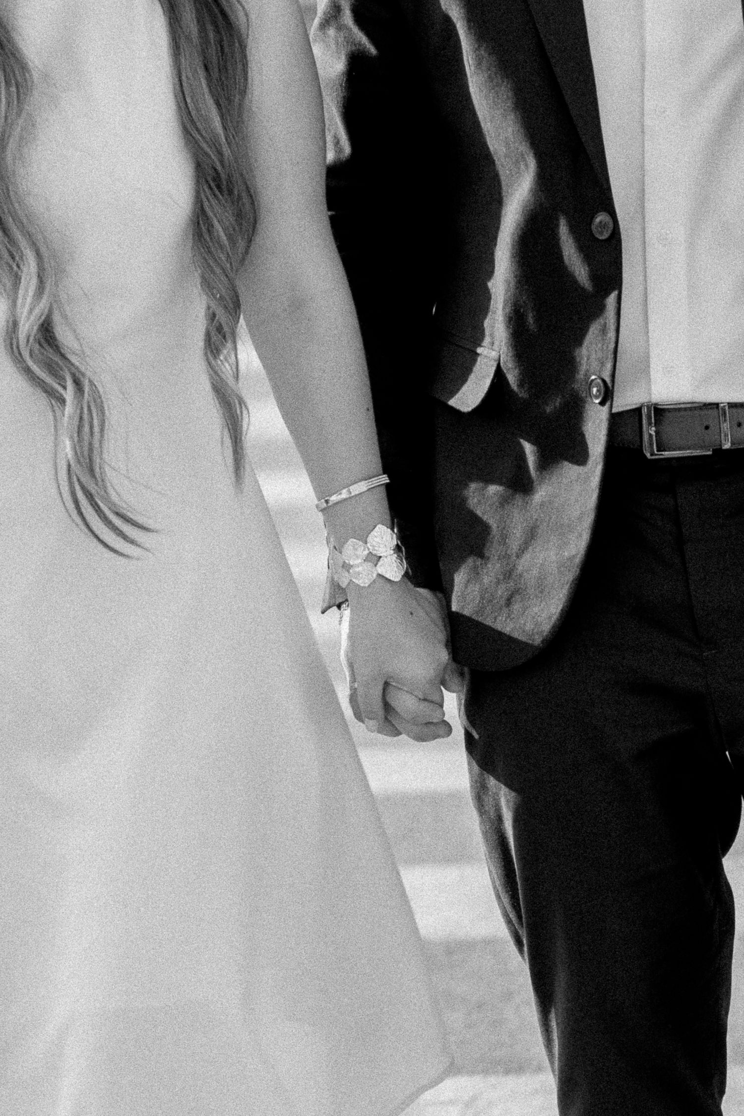 Black and white photo of a bride and groom holding hands, bride wearing a floral bracelet and white dress.