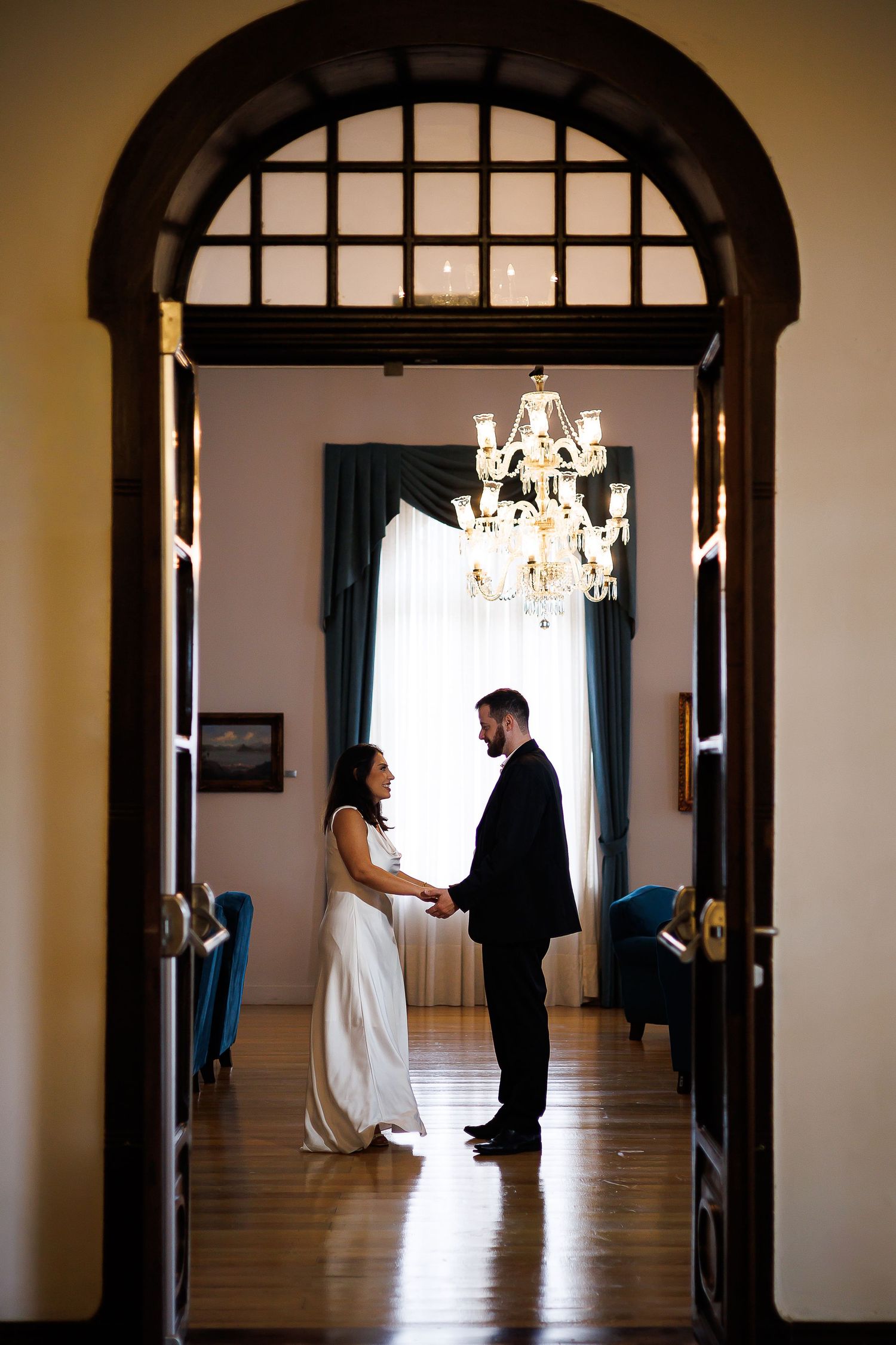 Bride and groom hold hands in elegant arched doorway with chandelier, sharing a romantic wedding moment indoors.