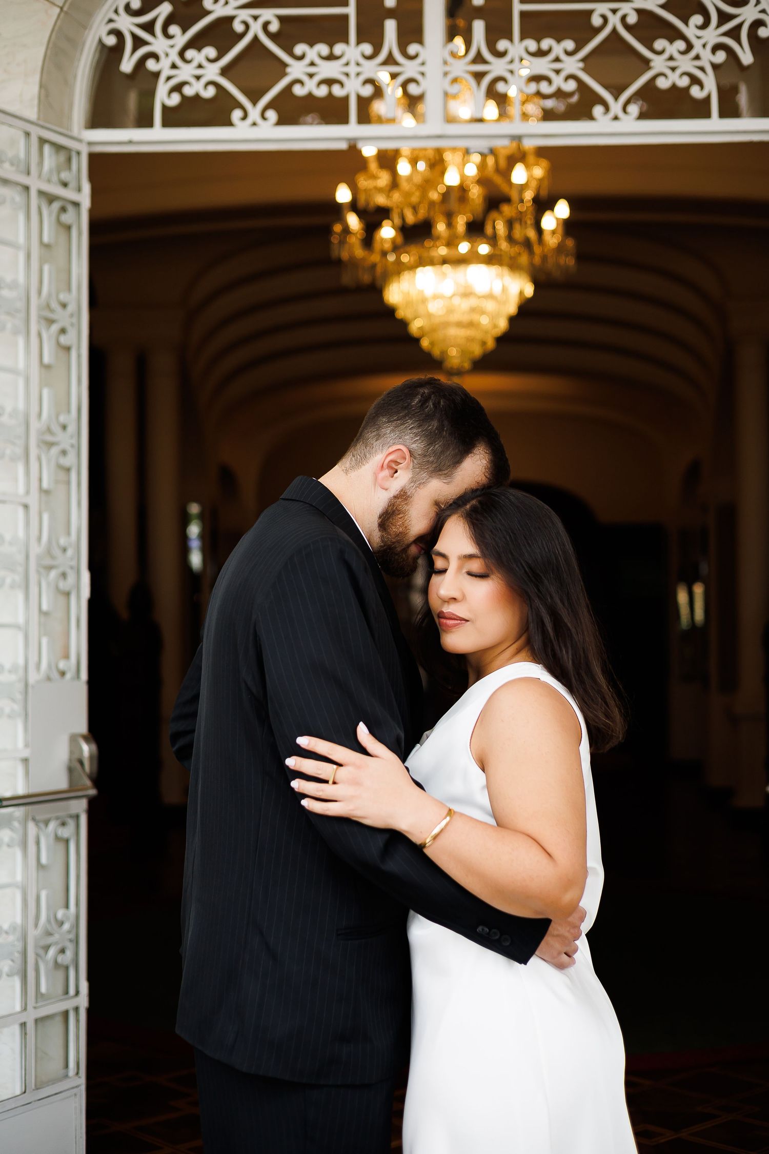 A couple in wedding attire embraces tenderly beneath a golden chandelier in an elegant venue doorway.