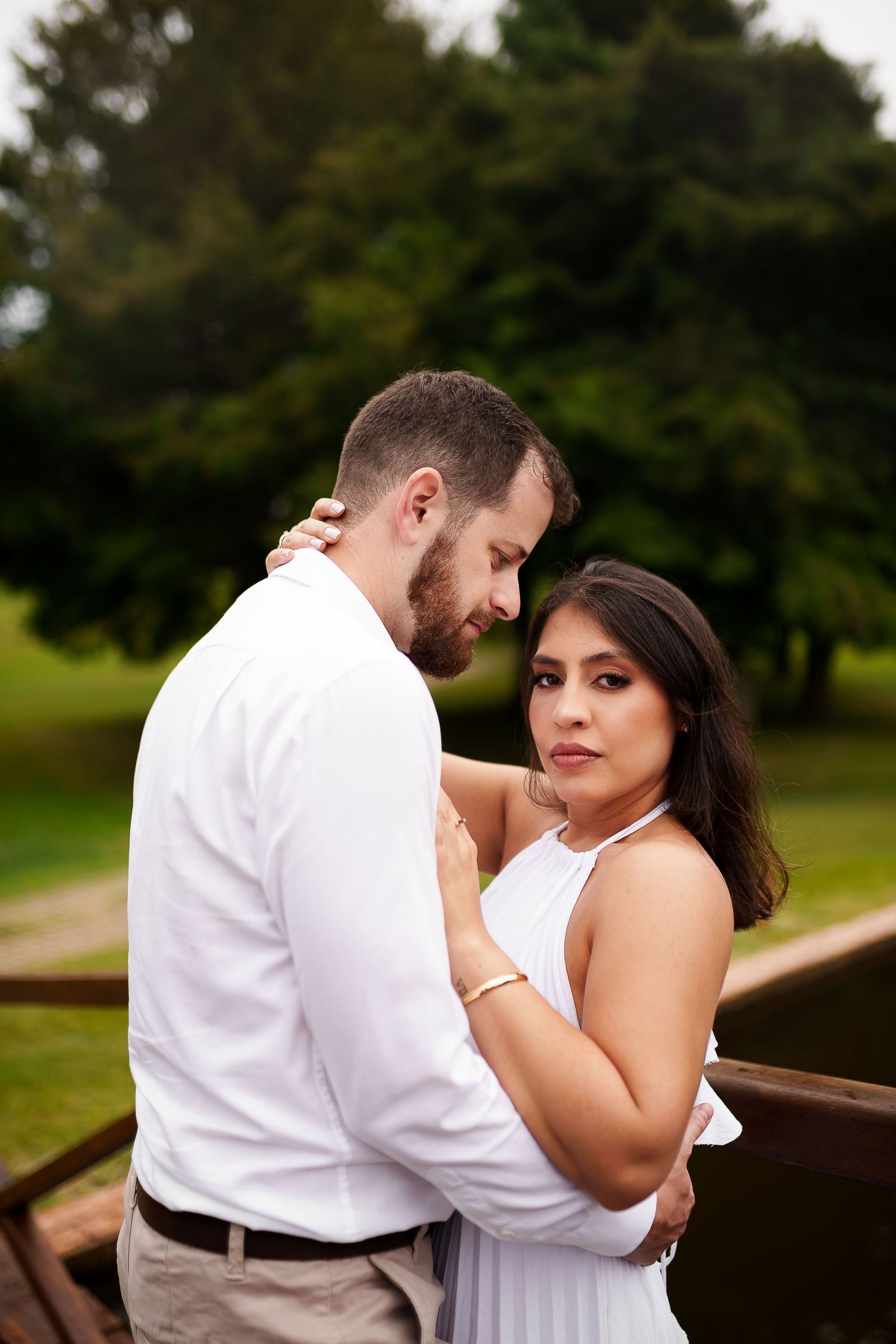 Couple in white outfits embracing on a wooden bridge surrounded by lush green trees in a romantic outdoor setting.