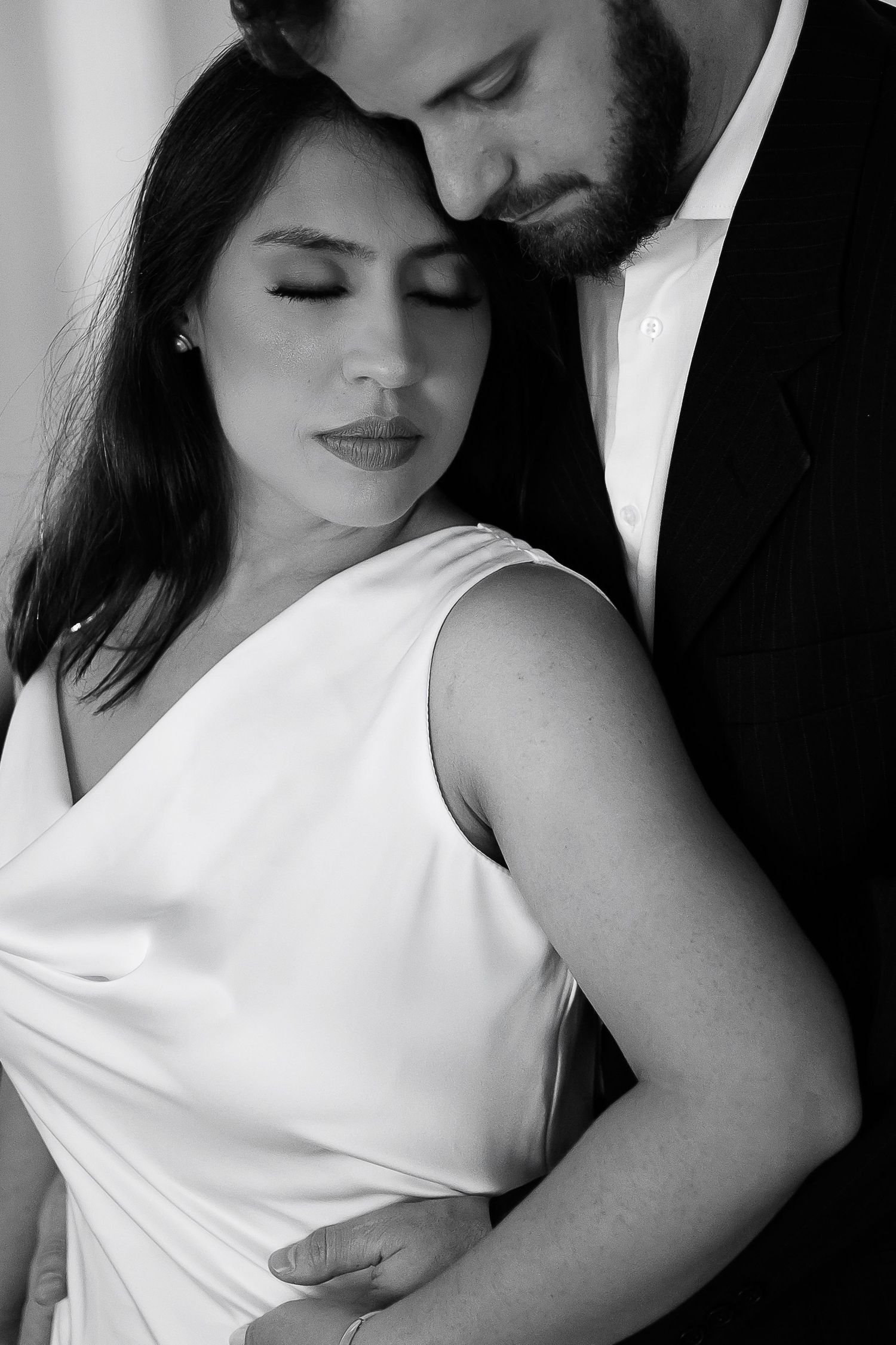Black and white photo of an elegant couple embracing, bride in white dress with groom in suit, eyes closed in tender moment.