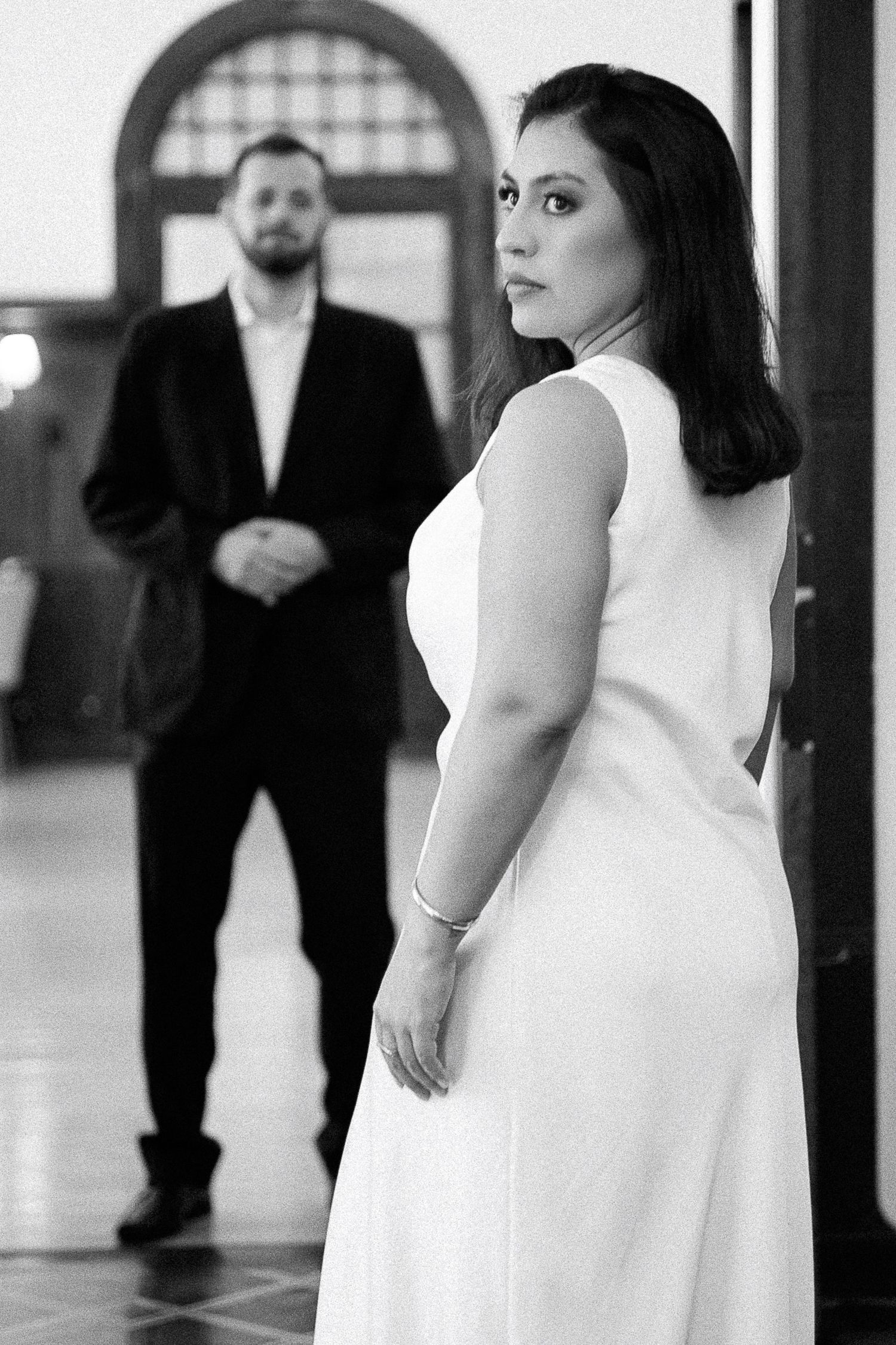 Black and white photo of bride in white dress looking back at groom in suit standing in elegant hall.