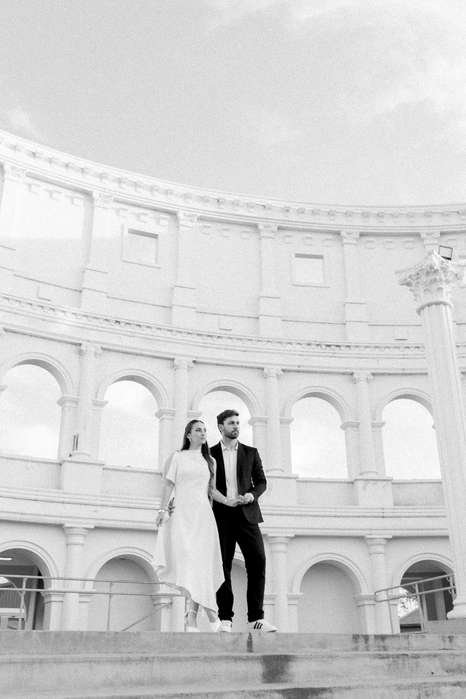 Black and white photo of an elegant couple standing on steps before a grand Roman colosseum-style architecture.