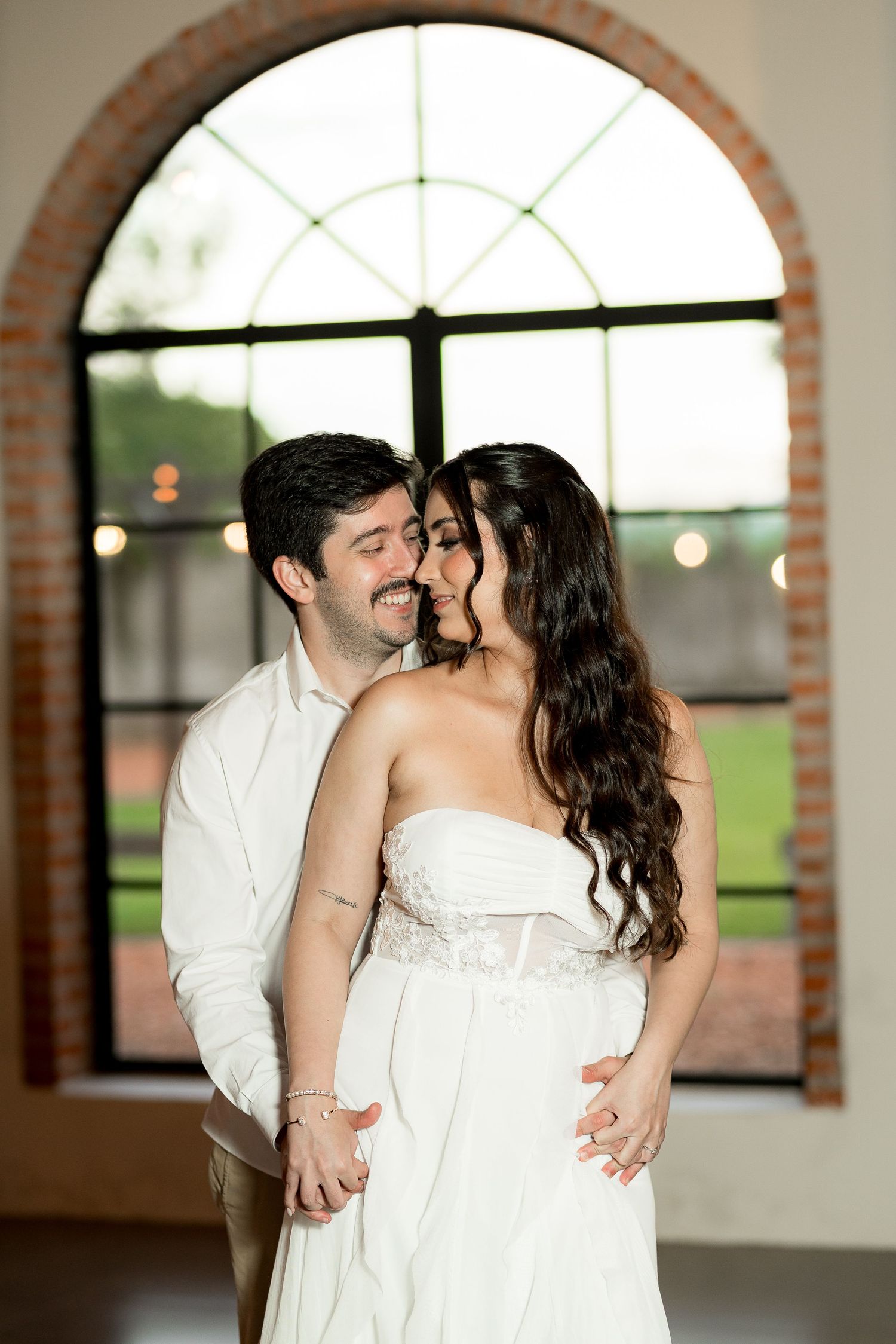A couple shares a romantic embrace in front of an arched window, the bride in a strapless white gown and groom in a white shirt.