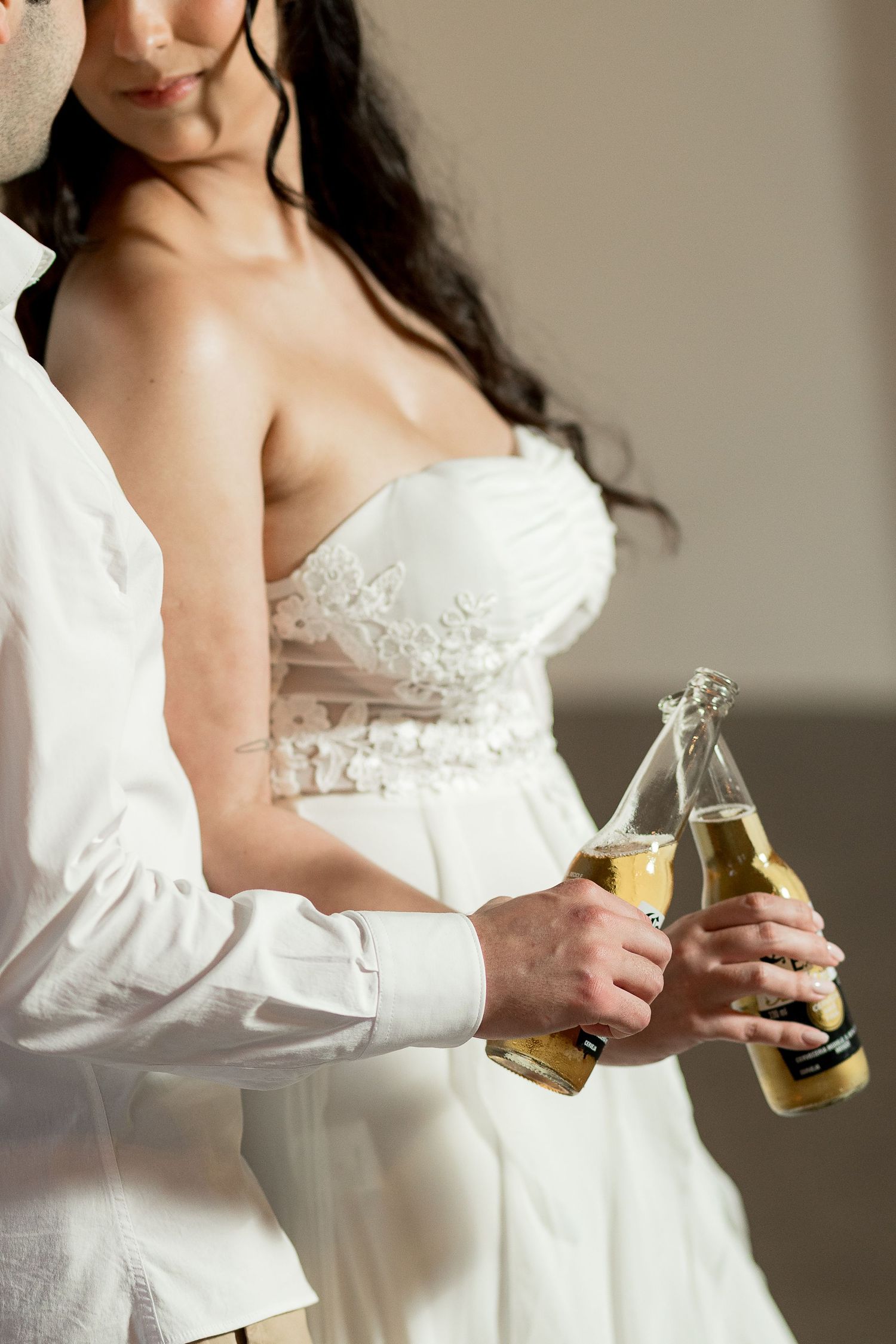 Bride and groom in white attire clinking beer bottles together during their wedding celebration.
