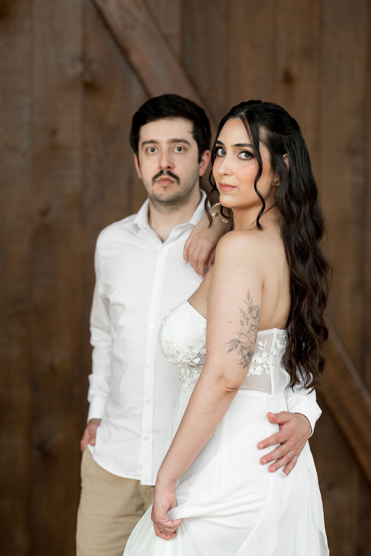 A bride in a white strapless gown with floral tattoo stands with a groom in white shirt against rustic wood backdrop.