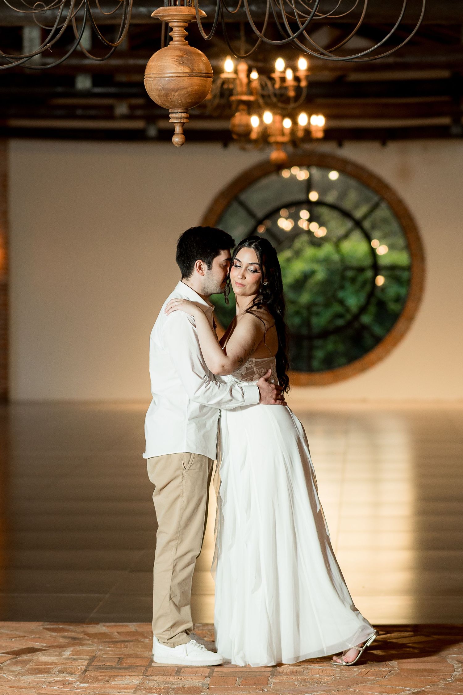 Couple sharing a romantic embrace in an elegant venue with chandelier and round window backdrop.