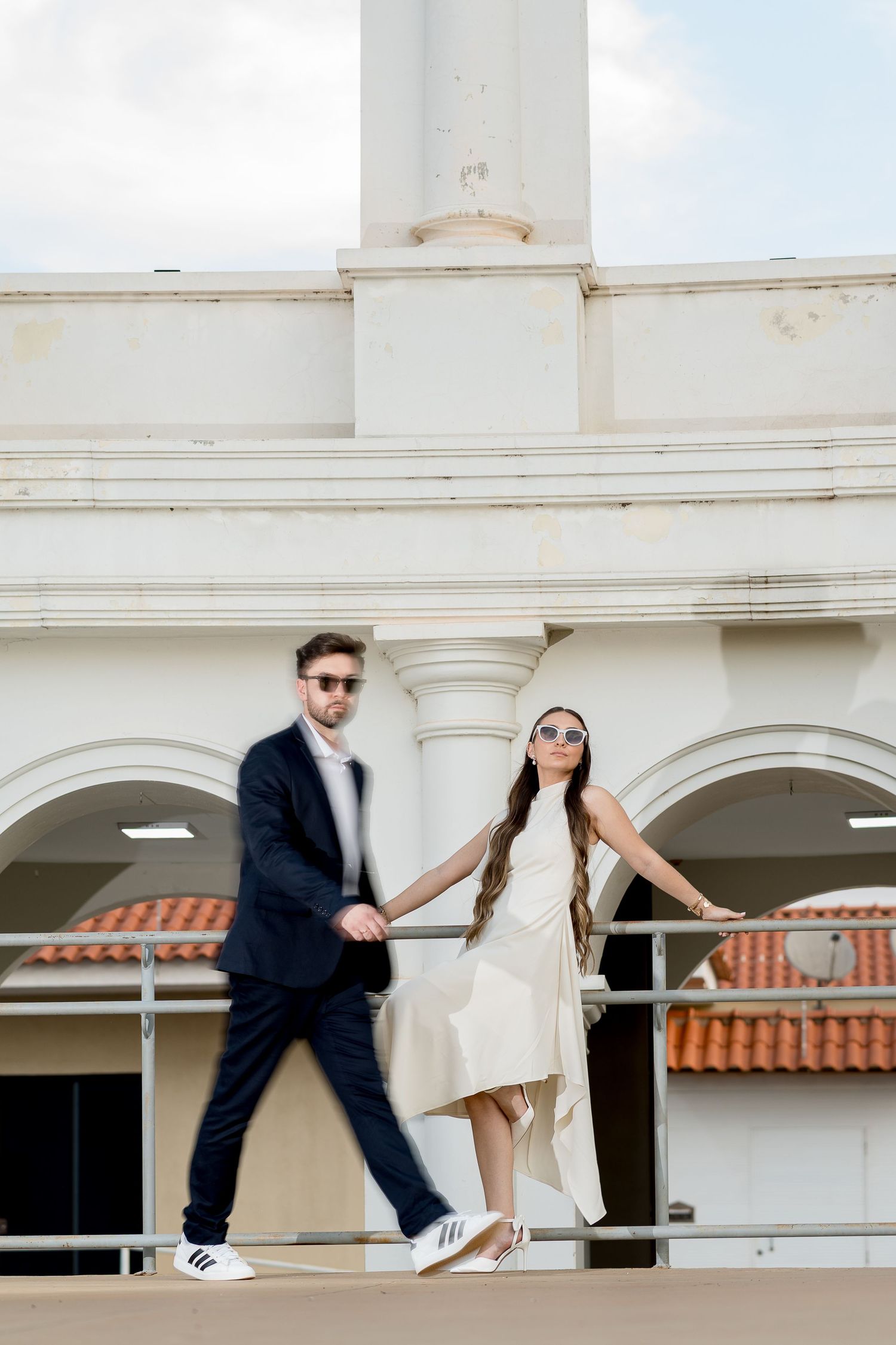 Stylish couple holding hands on a balcony, man in navy suit and woman in cream dress with sunglasses, white building backdrop.
