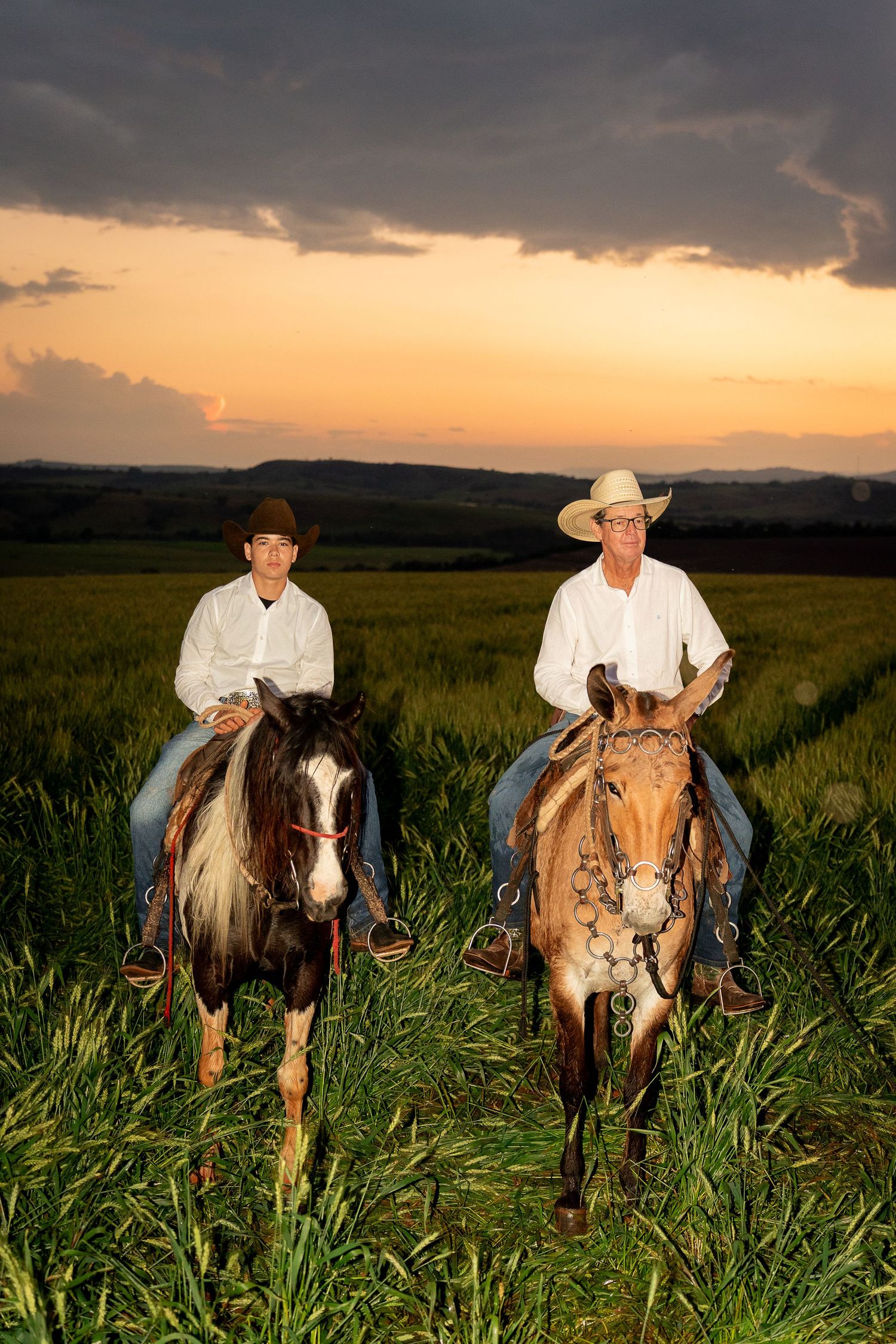 Two cowboys in white shirts ride horses through a lush green field at sunset with dramatic cloudy skies.