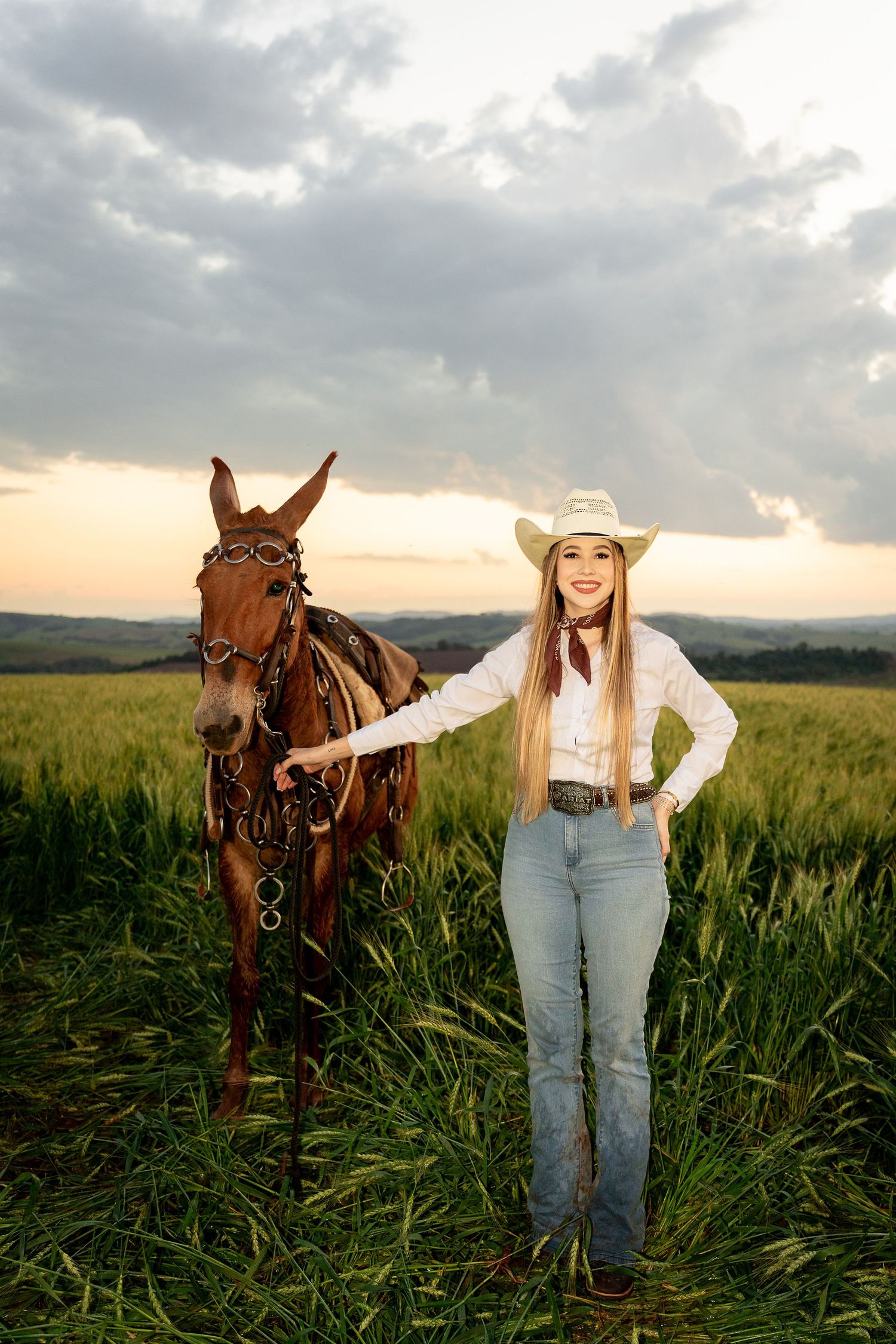 Young woman in cowboy hat and jeans stands smiling beside a brown horse in a green field at sunset.