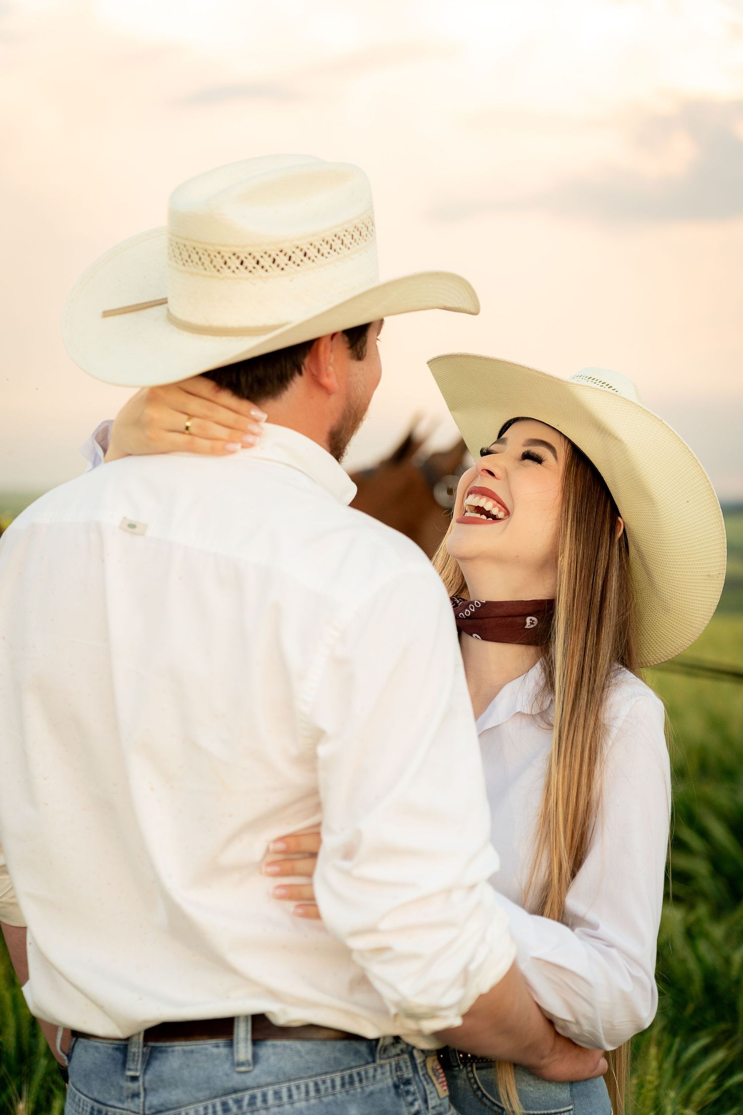 A smiling couple in white shirts and cowboy hats embrace in a green field at sunset, with a horse visible behind them.