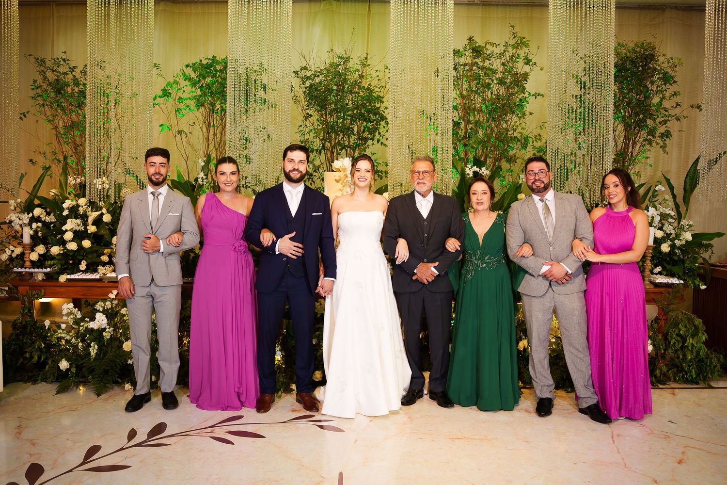 Wedding party poses together in front of lush greenery backdrop, bride in white gown flanked by guests in pink and green formal attire.