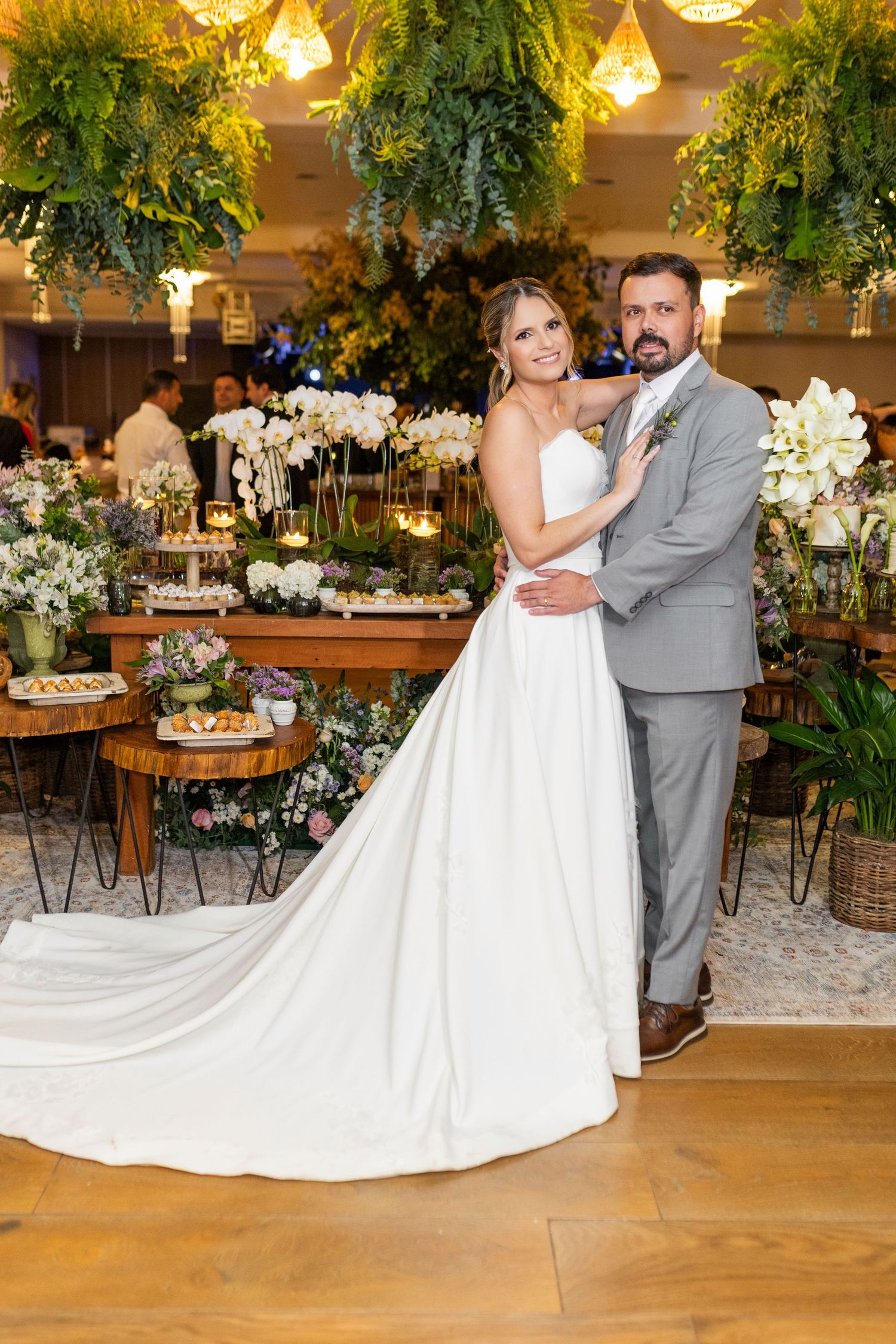 Bride in white gown and groom in gray suit pose together at elegant floral wedding reception venue.