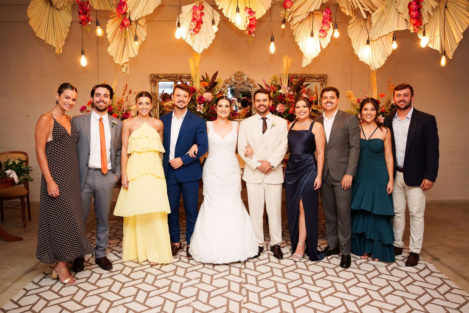 A joyful wedding group photo featuring the bride and groom surrounded by elegantly dressed guests under decorative floral ceiling.