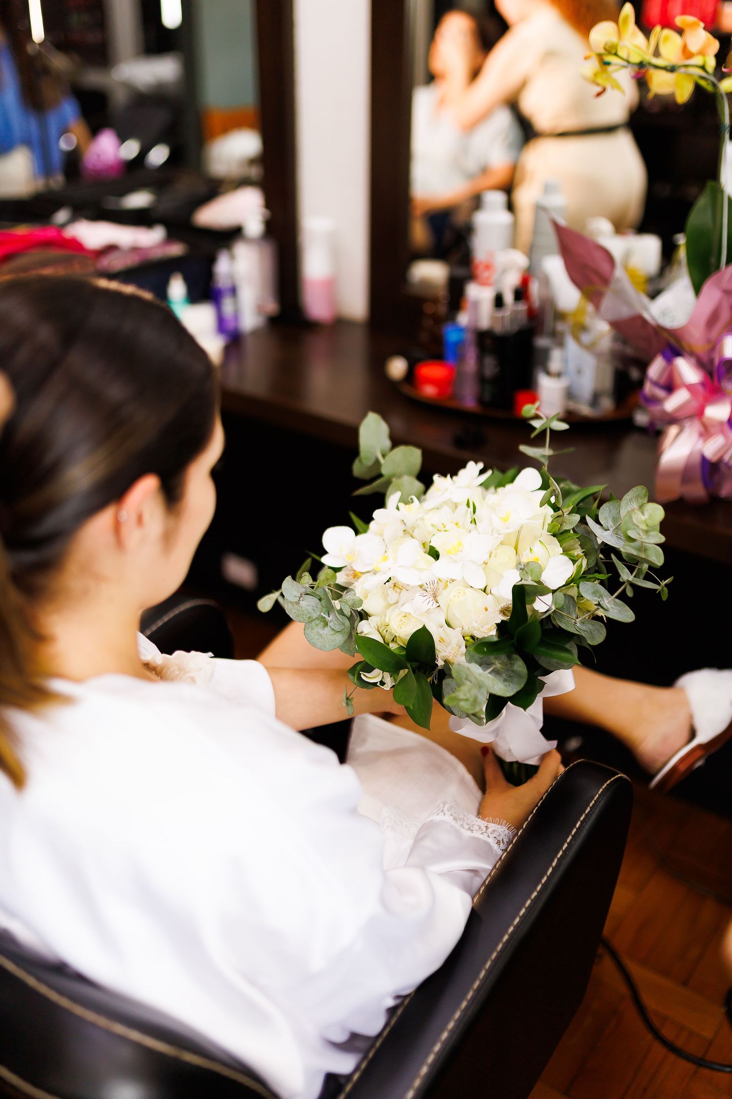 Bride in white robe holding elegant bouquet of white flowers at salon mirror during wedding preparation.