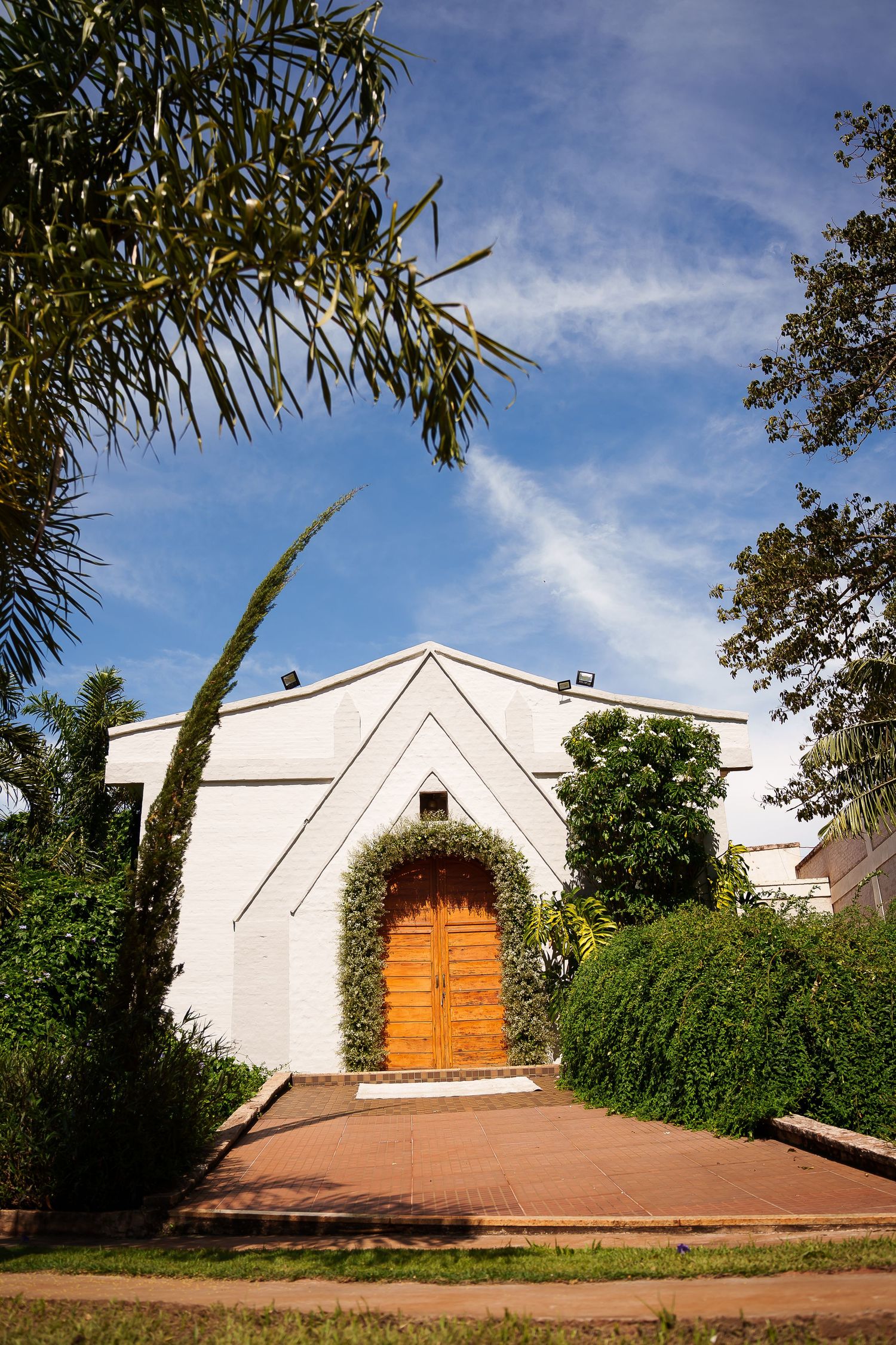White stucco building with arched wooden door, framed by lush greenery and palm trees under a blue sky.