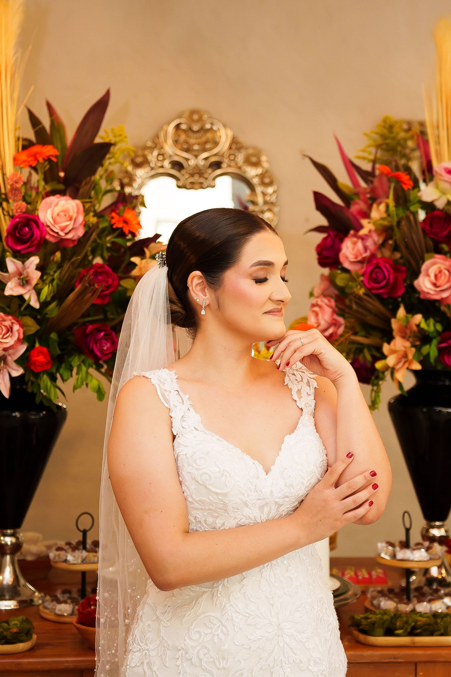 Bride in white lace gown and veil poses gracefully before lush floral arrangements of roses and tropical blooms.