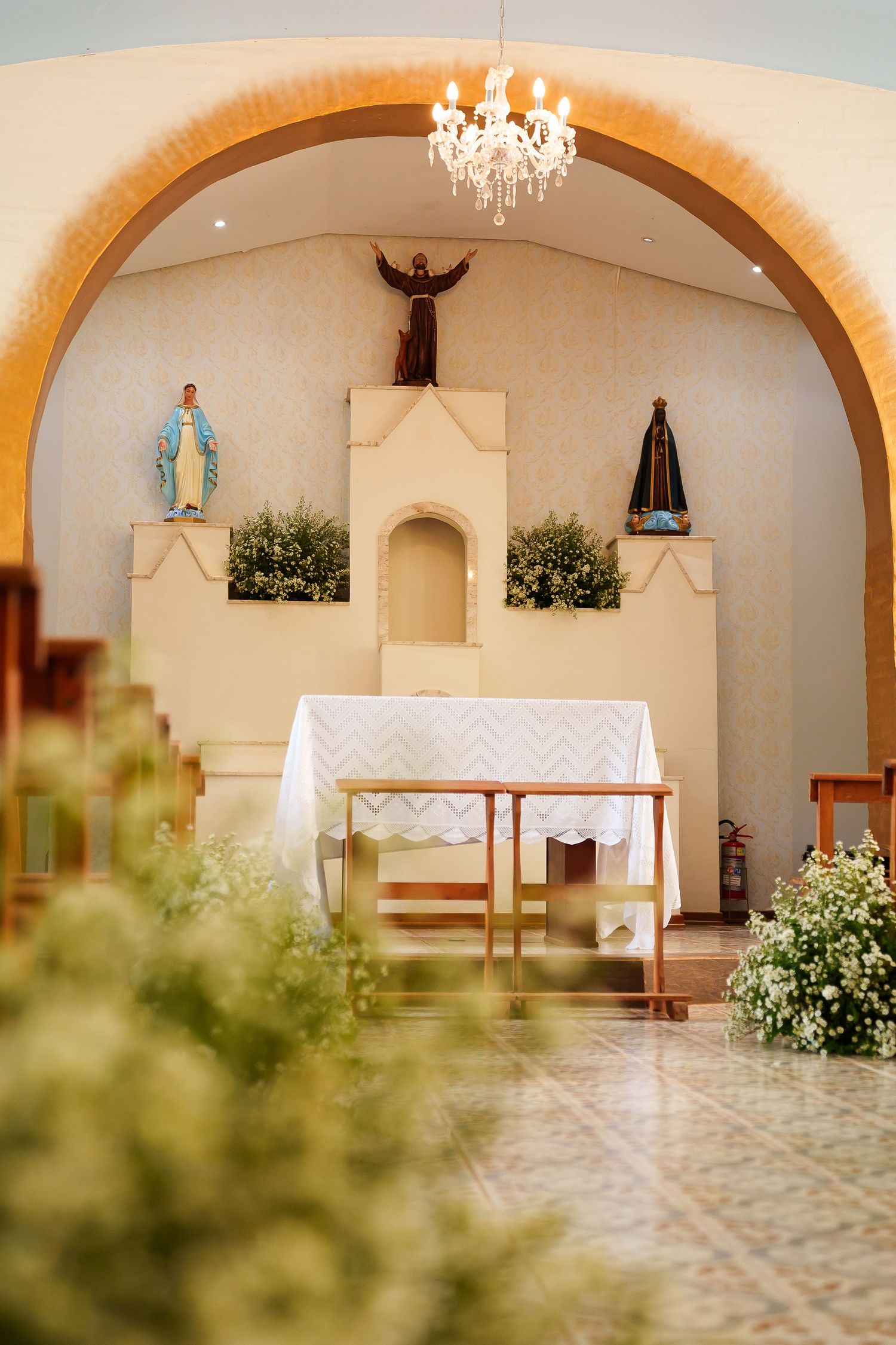 Small chapel interior with altar, religious statues, chandelier, white flowers, and wooden pews under an arched entrance.