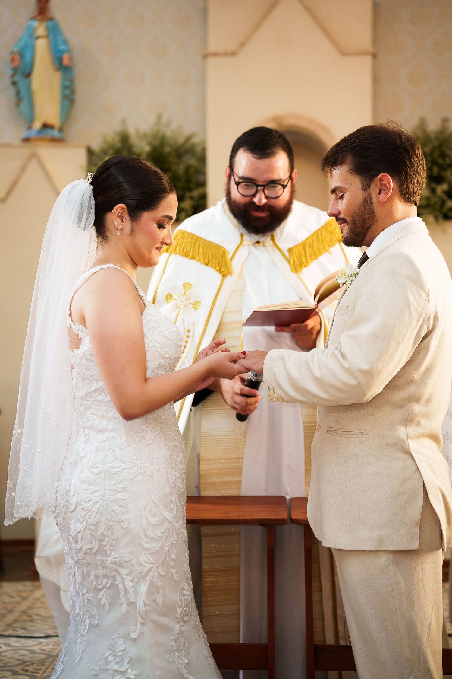 Bride and groom exchange rings at church altar with bearded priest in white and gold vestments during wedding ceremony.