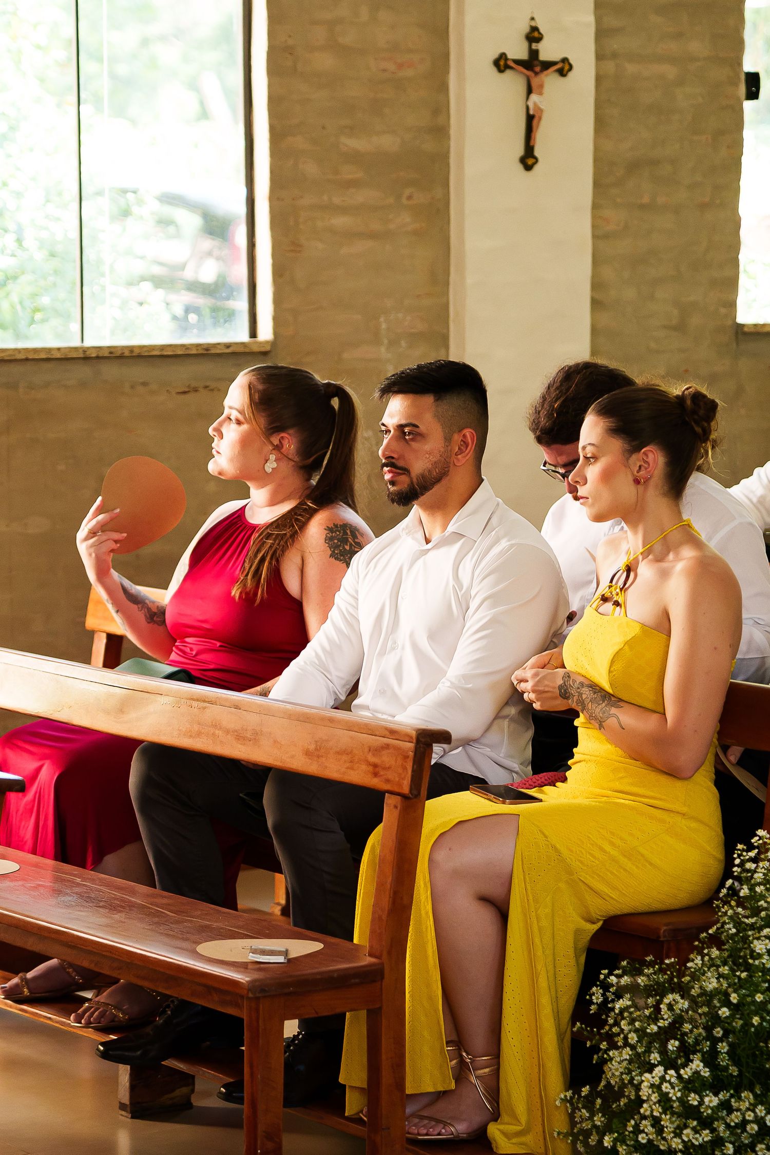 Guests seated in church pews during a wedding ceremony, wearing elegant red and yellow dresses and white shirt.
