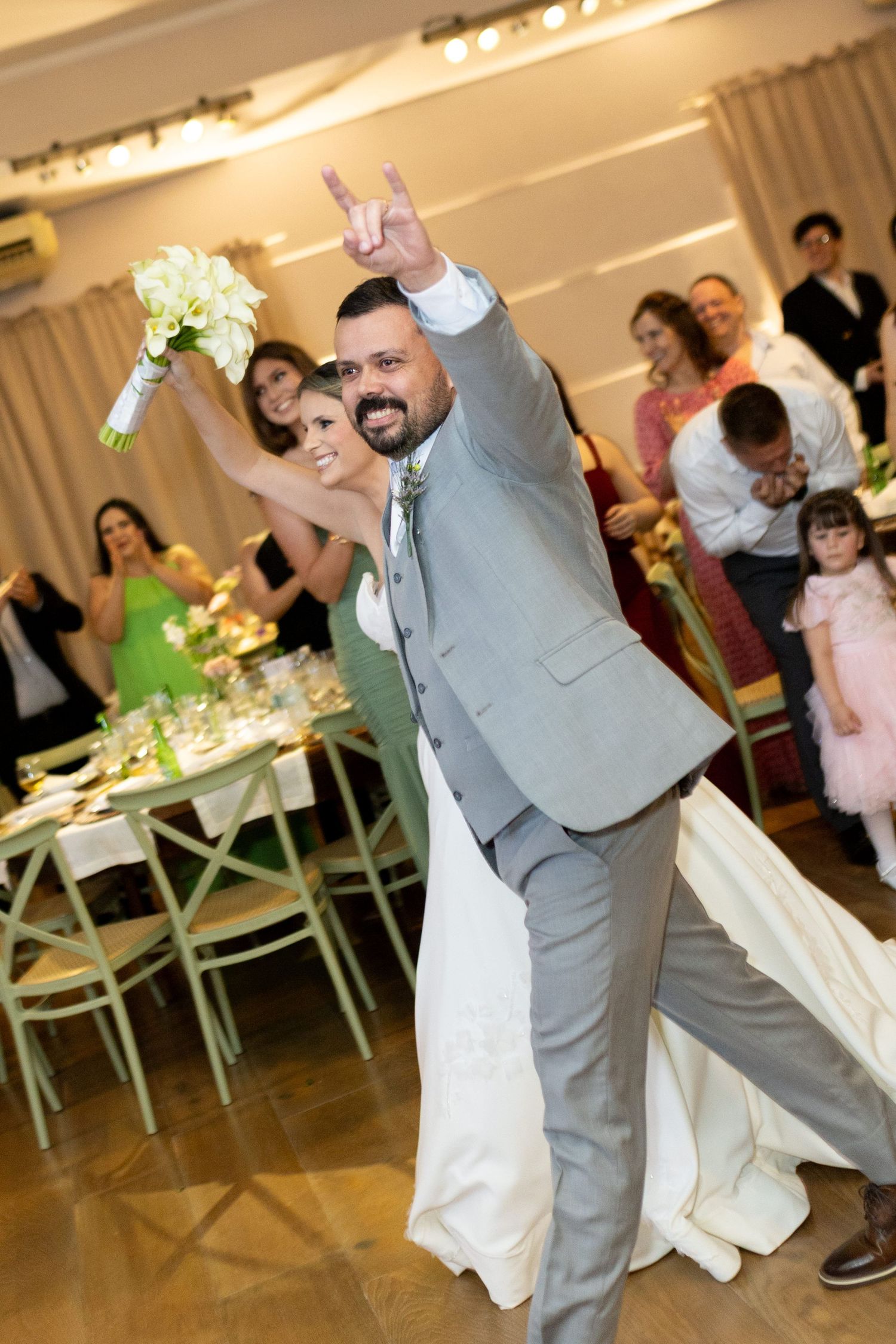 Bride and groom make grand entrance at wedding reception, groom gesturing rock sign while bride holds white bouquet.