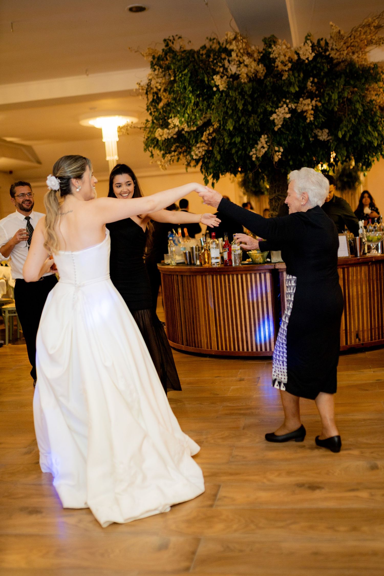 Bride in white gown dancing joyfully with elderly woman in black dress at wedding reception.