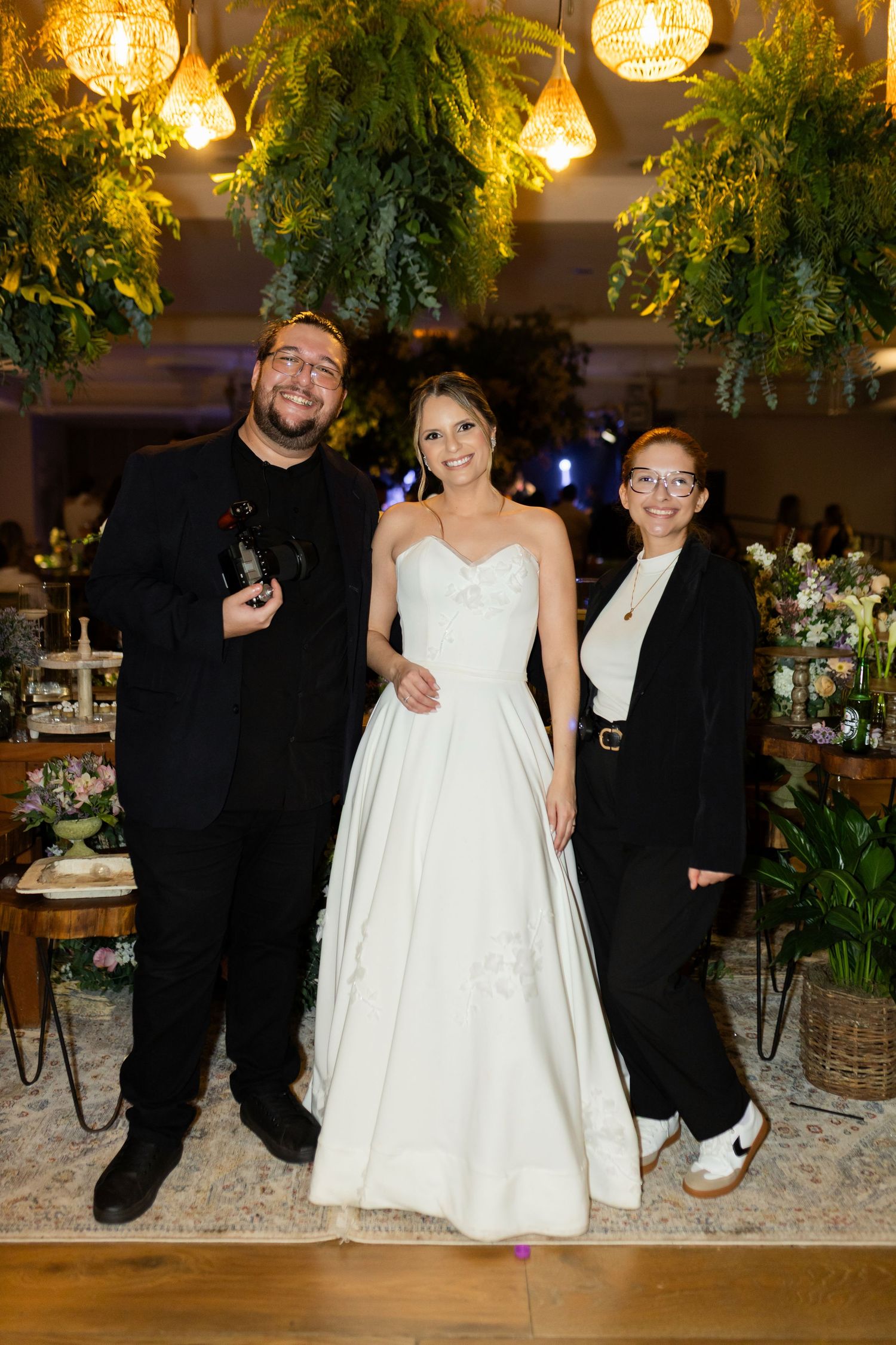 Bride in white strapless gown poses with two guests in black attire at an elegant floral wedding reception.