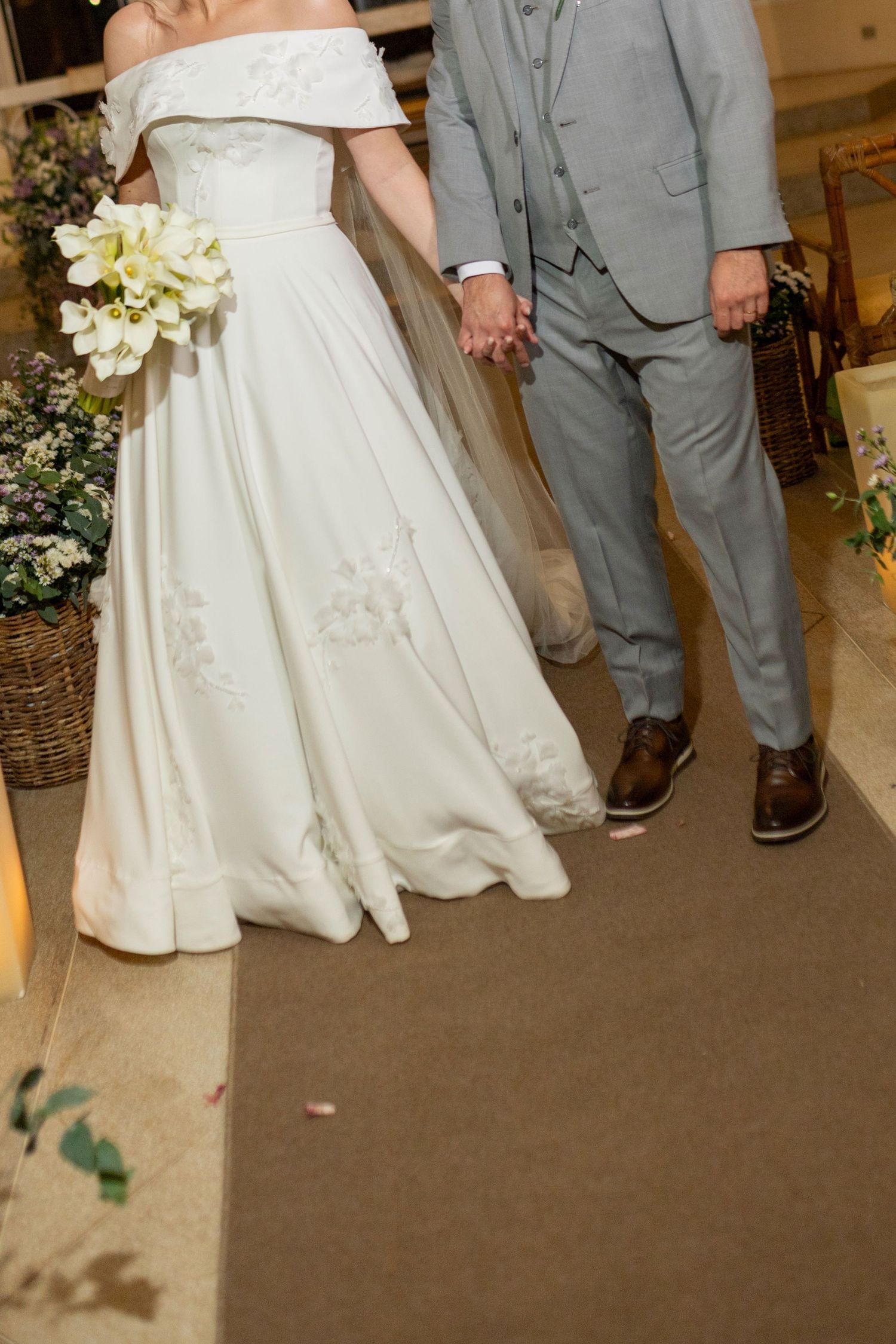 Bride in off-shoulder white gown holding bouquet, groom in gray suit, holding hands at wedding ceremony.