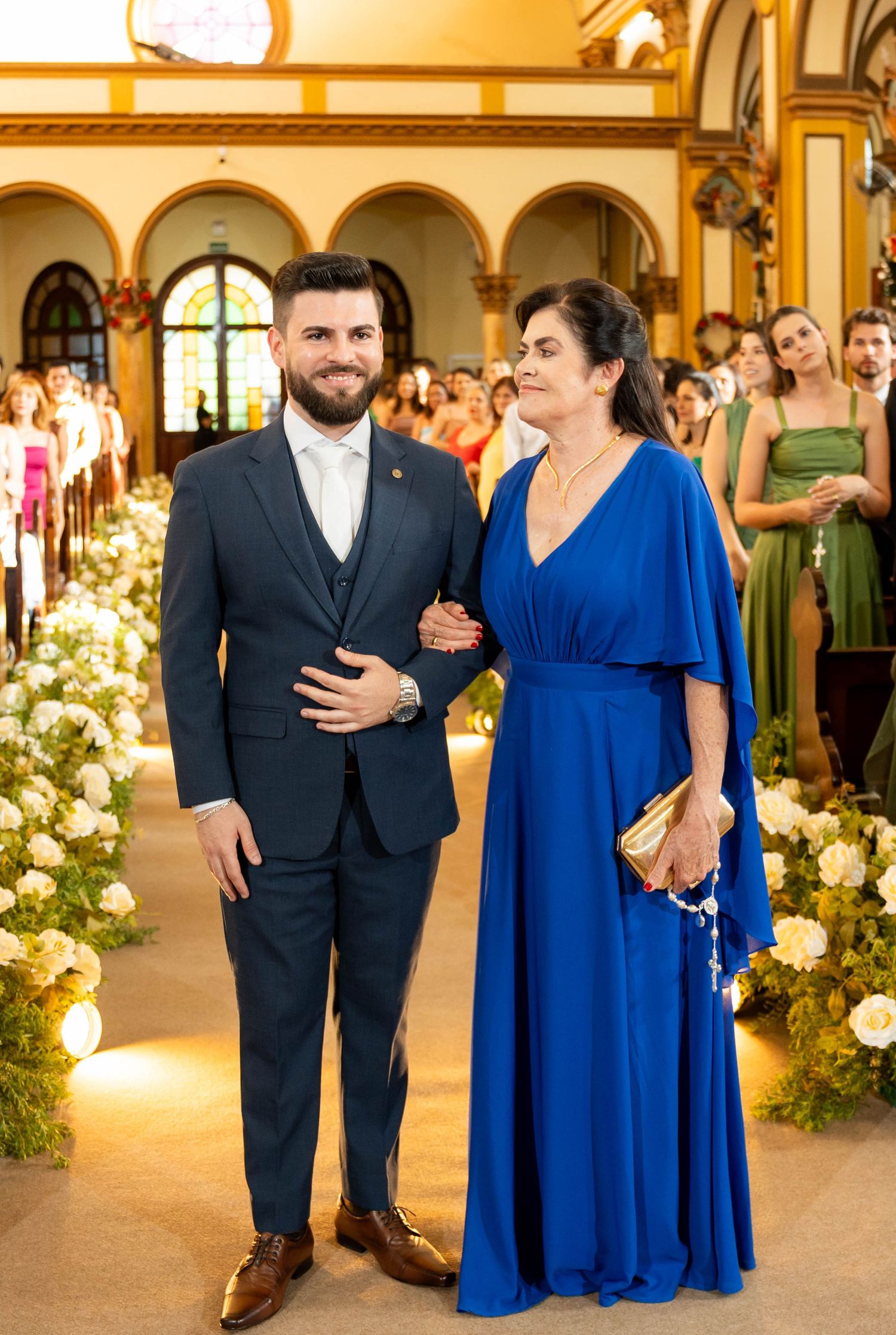 A man in a navy suit and a woman in a royal blue gown walk down a floral-decorated church aisle at a wedding.