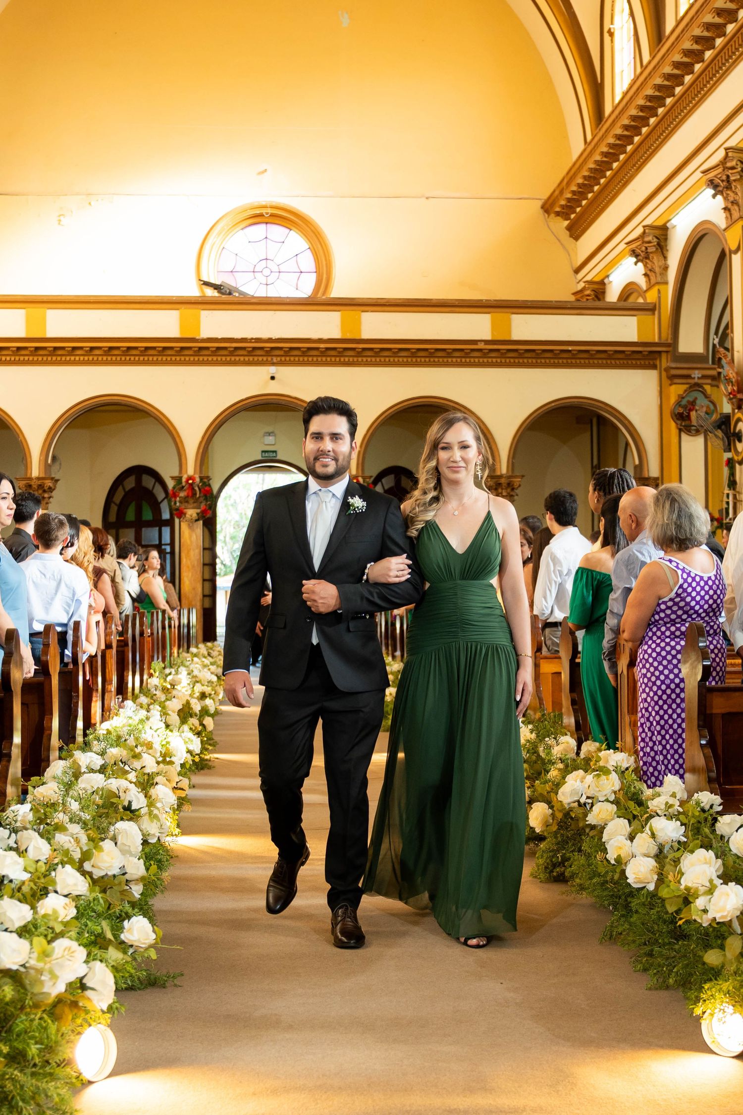 A couple walks down a church aisle lined with white flowers, man in black suit, woman in emerald green dress.