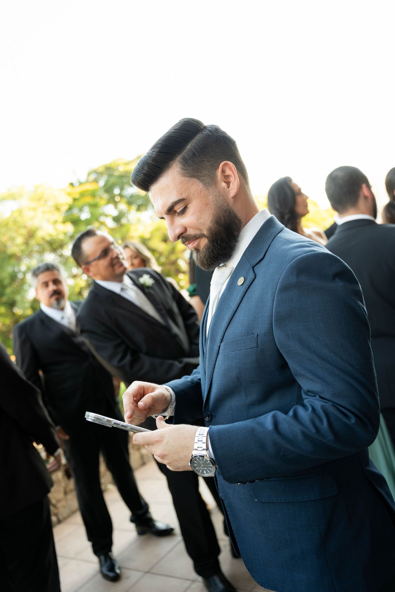 Bearded man in blue suit checks phone at outdoor formal event with guests in background.