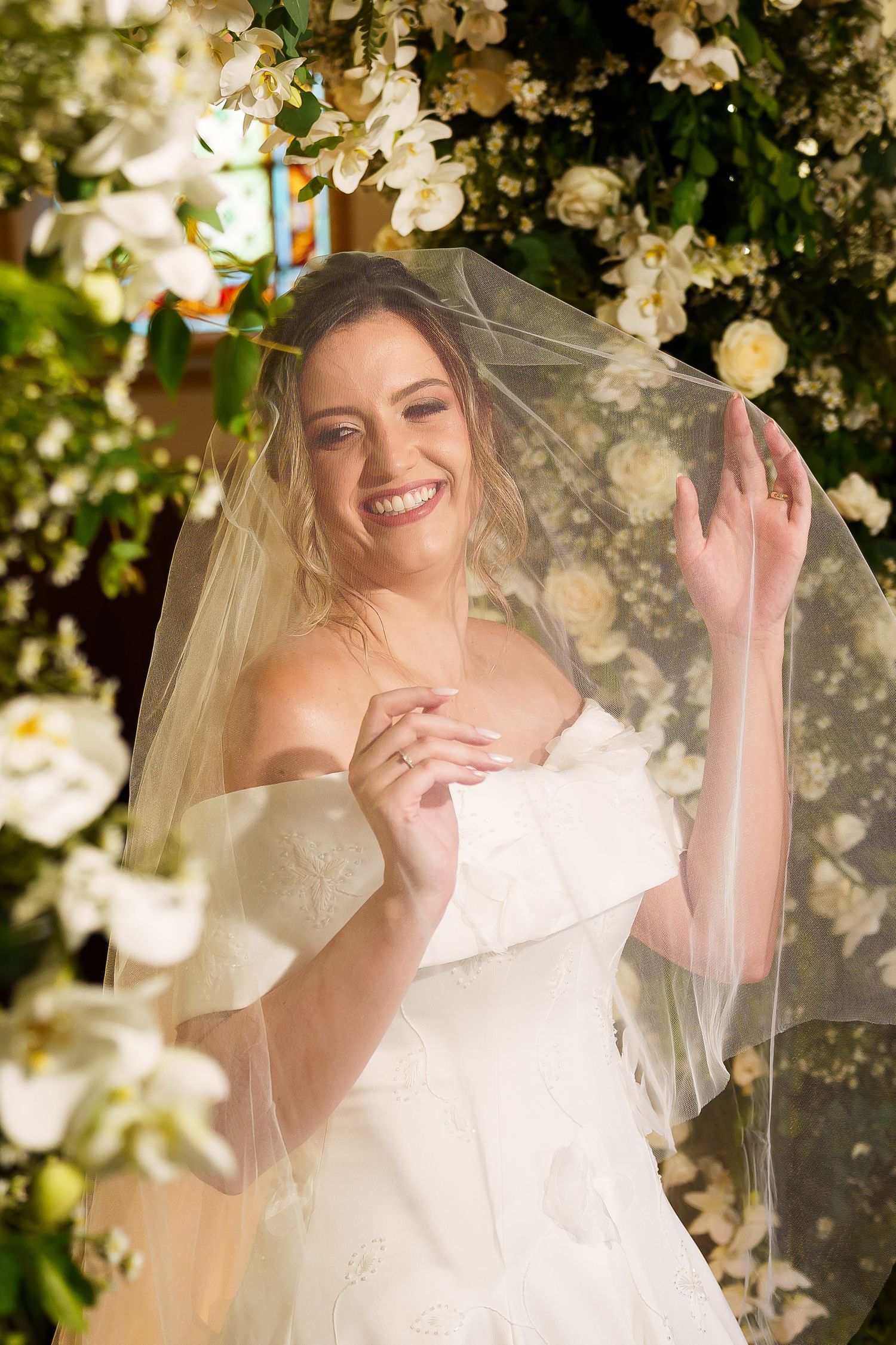 Smiling bride in white off-shoulder gown and veil poses among lush white floral arrangements at wedding ceremony.