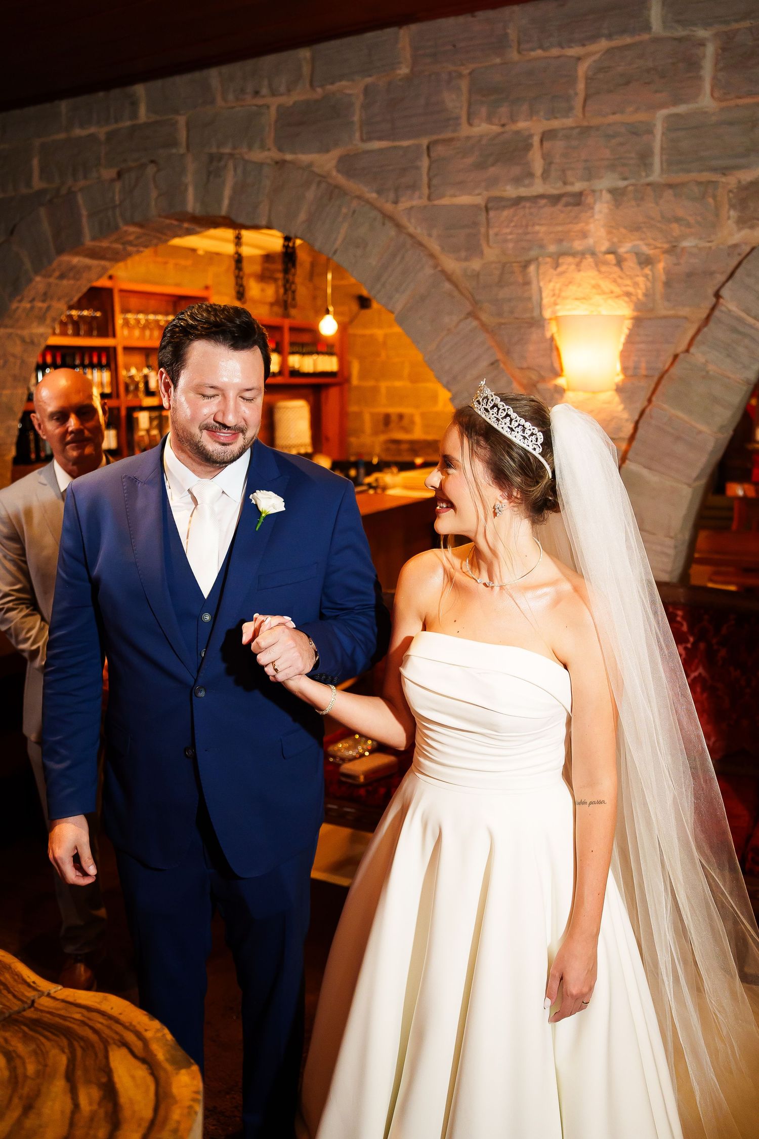 Bride in white gown and tiara holds groom's hand in blue suit, smiling in a warmly lit stone arch venue.
