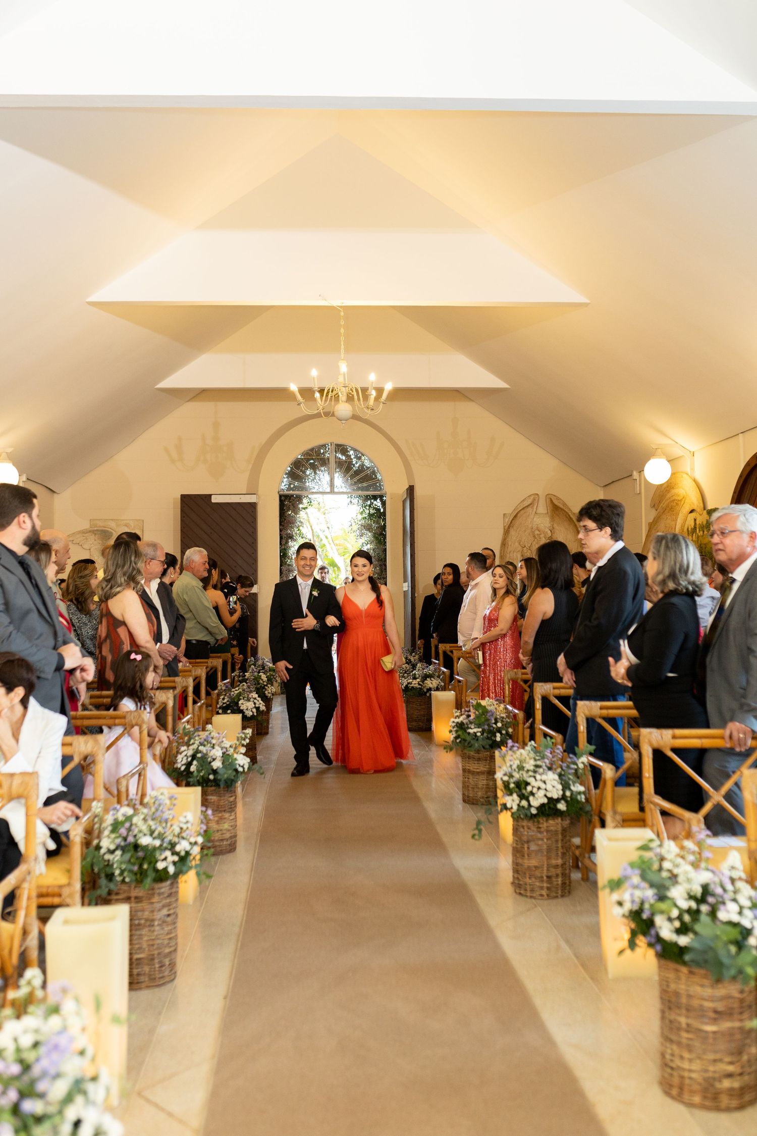 Bride in red gown walks down chapel aisle with escort, flanked by floral arrangements in wicker baskets.