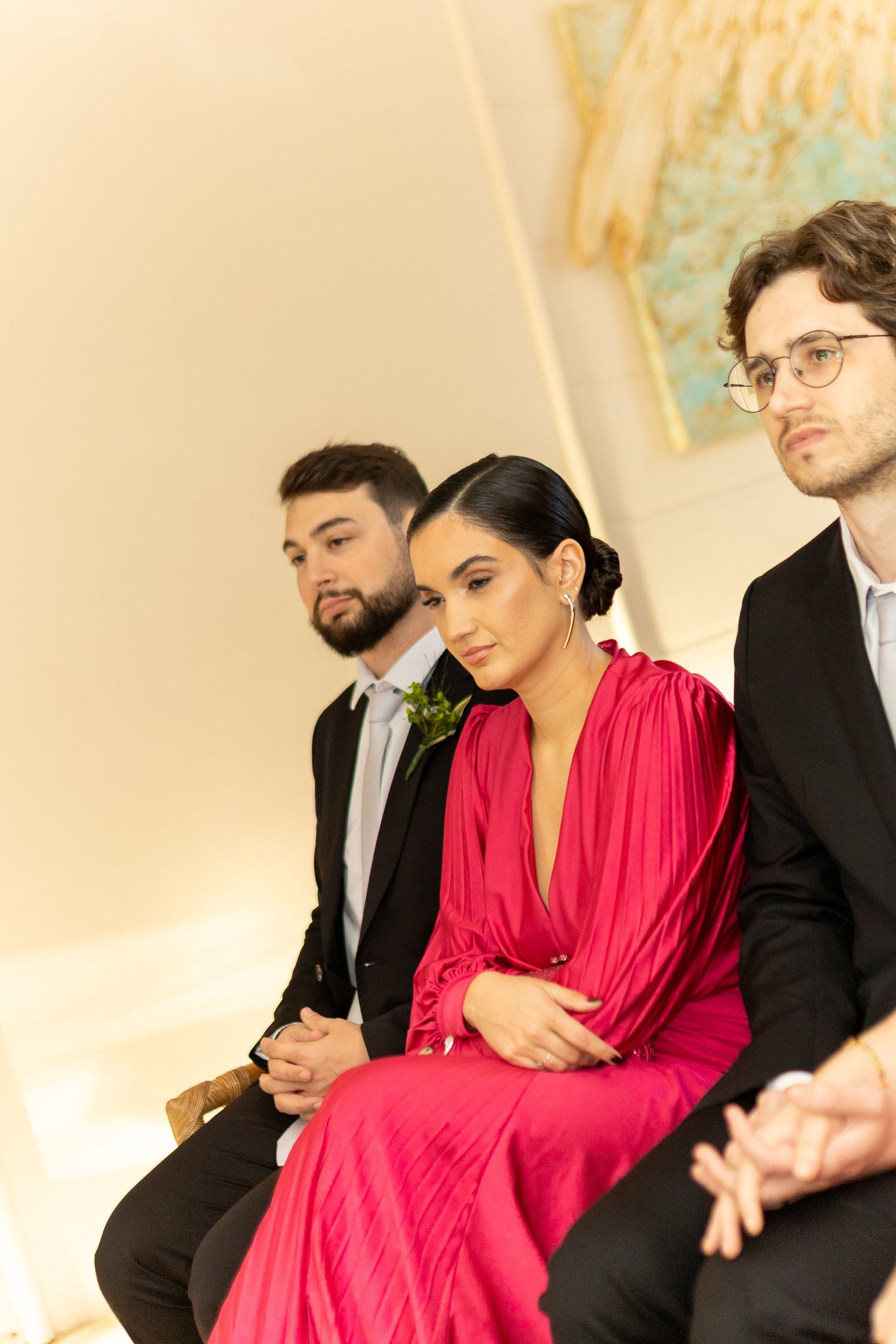 Three elegantly dressed guests seated at a formal event, woman in red gown flanked by men in black suits.